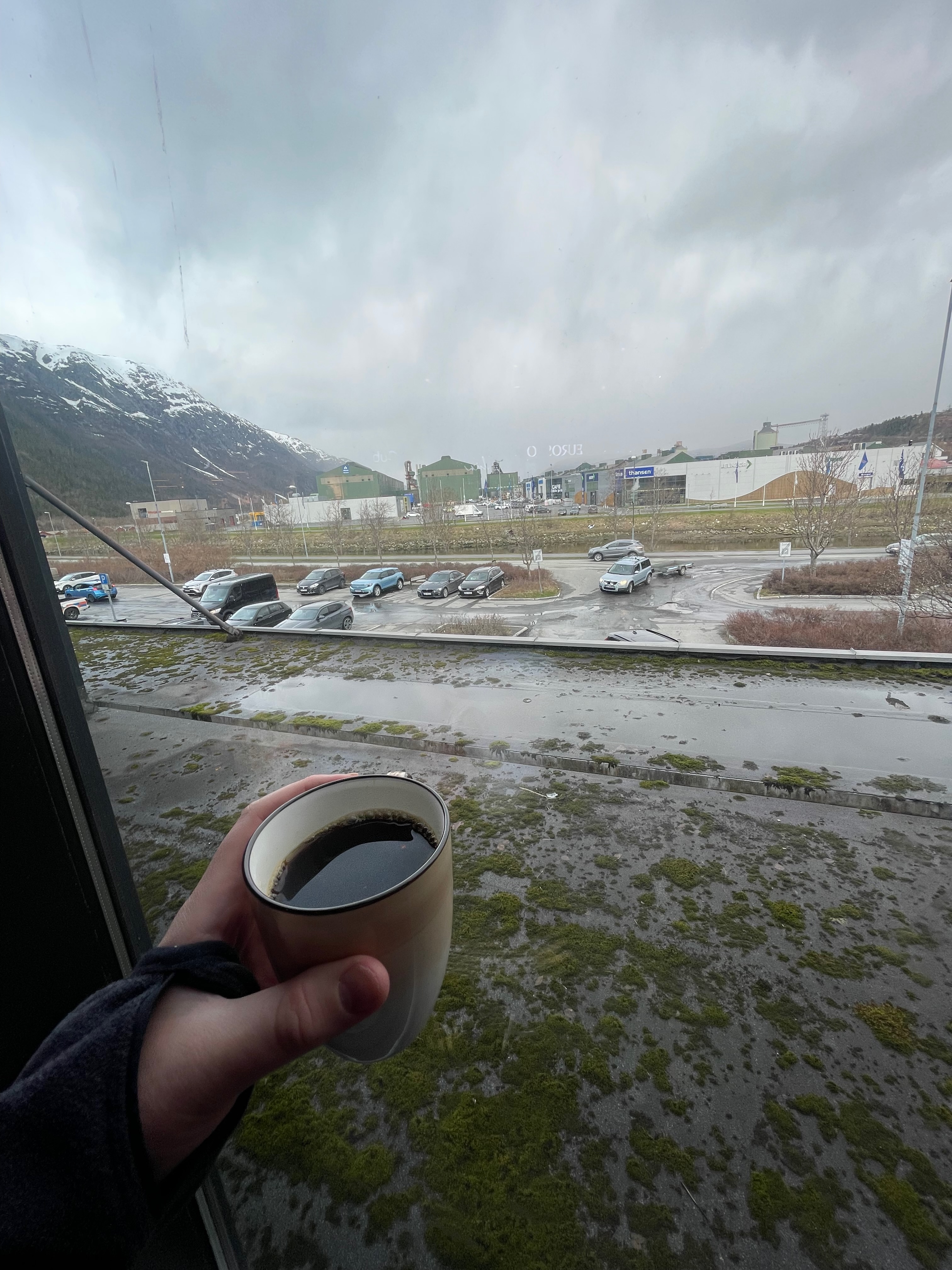 Cafe Sjøsiden coffee stop in Mosjøen, showing black coffee by the window with rainy weather, parking area, mossy roof, and mountains outside.