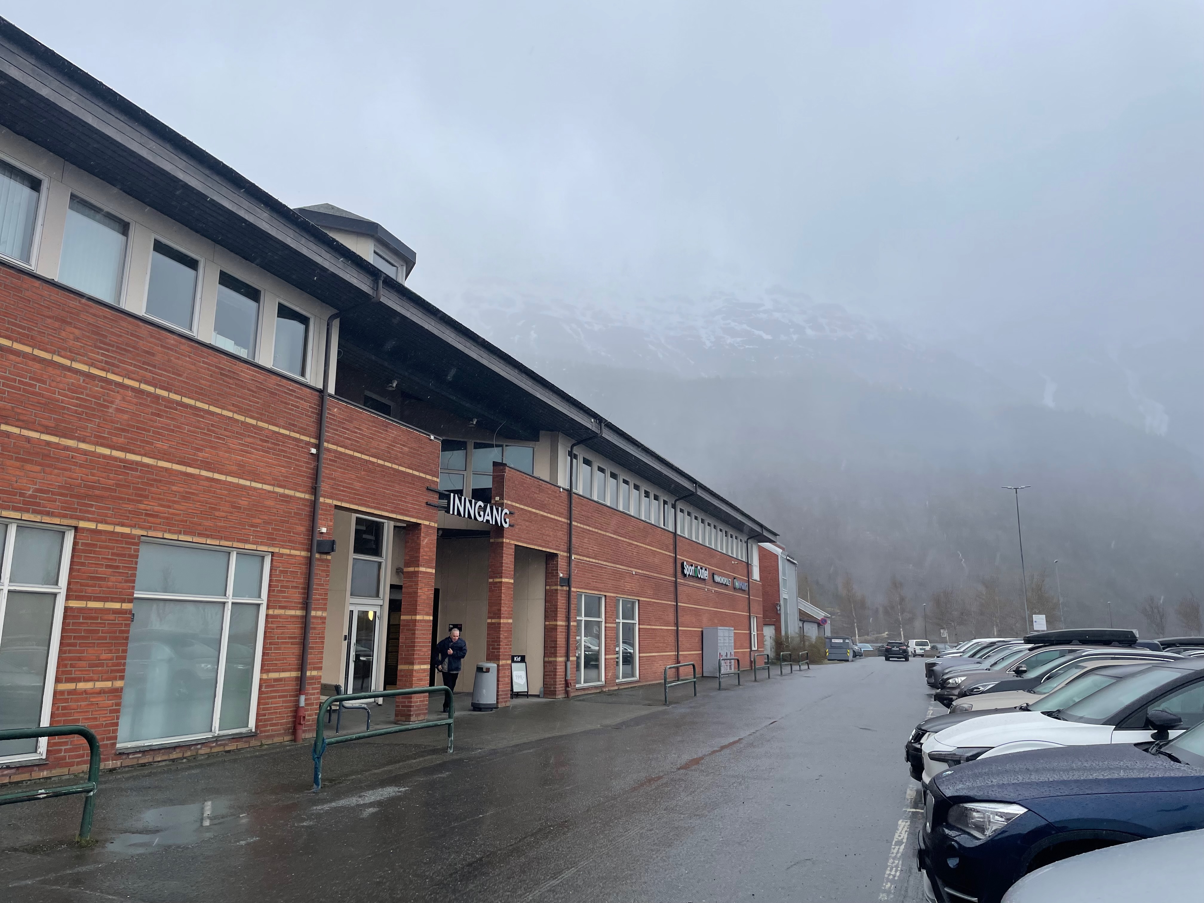 Cafe Sjøsiden in Mosjøen, exterior view of Alti Sjøsiden shopping center with a rainy parking lot and misty mountains during a Norway road trip stop.