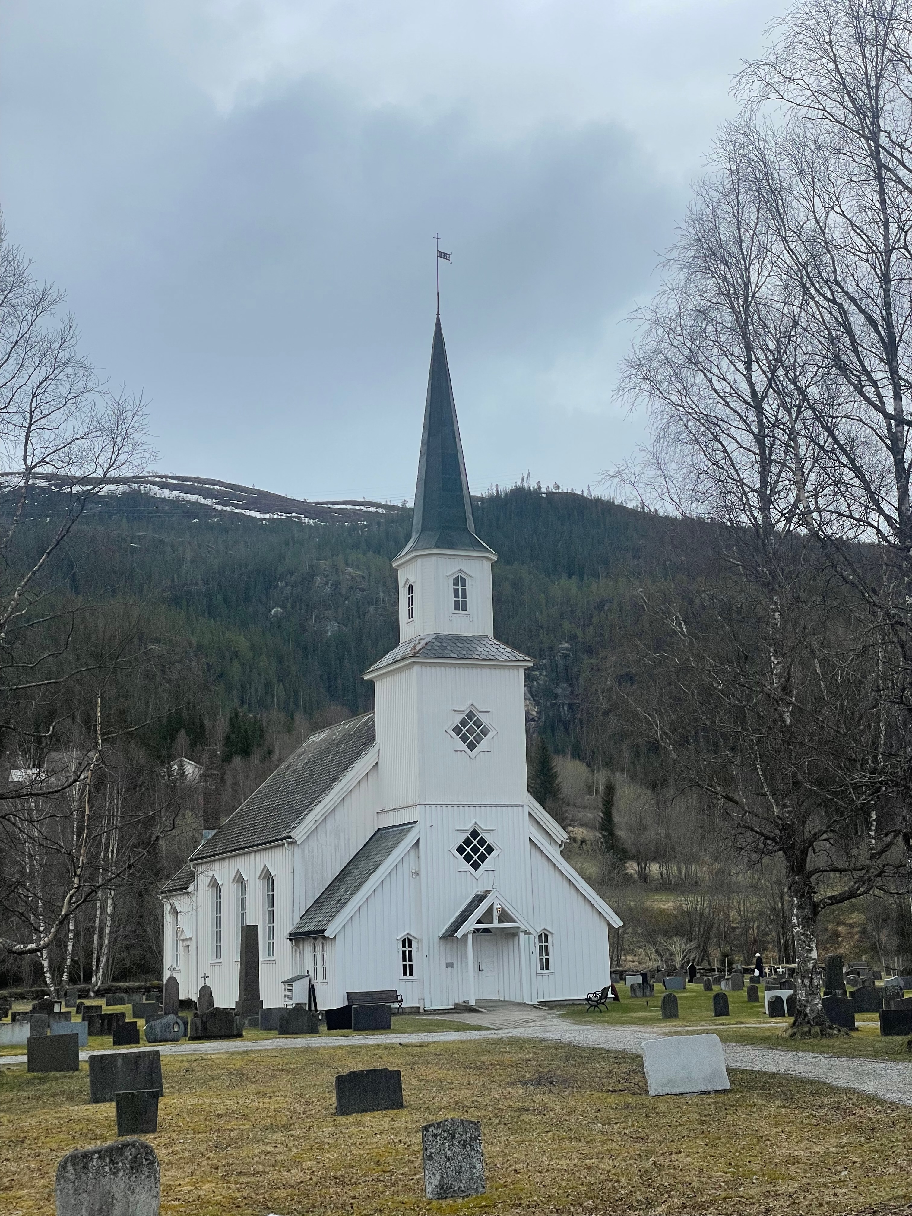 Grane Church in Grane near Trofors, a white wooden church with a tall dark steeple, surrounded by gravestones, bare trees, and forested mountains in Northern Norway.