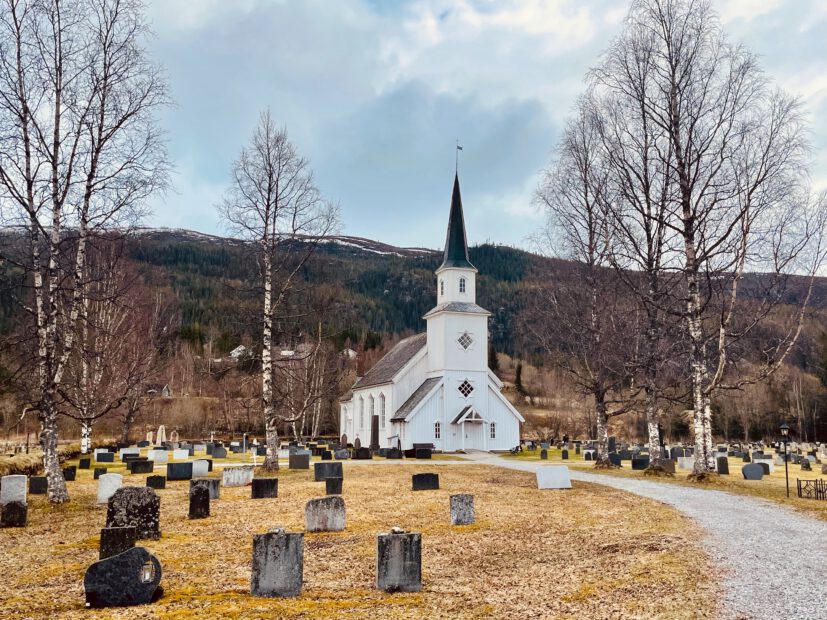 Grane Church in Grane near Trofors, a white wooden church surrounded by graves, birch trees, a gravel path, and mountain scenery in Northern Norway.