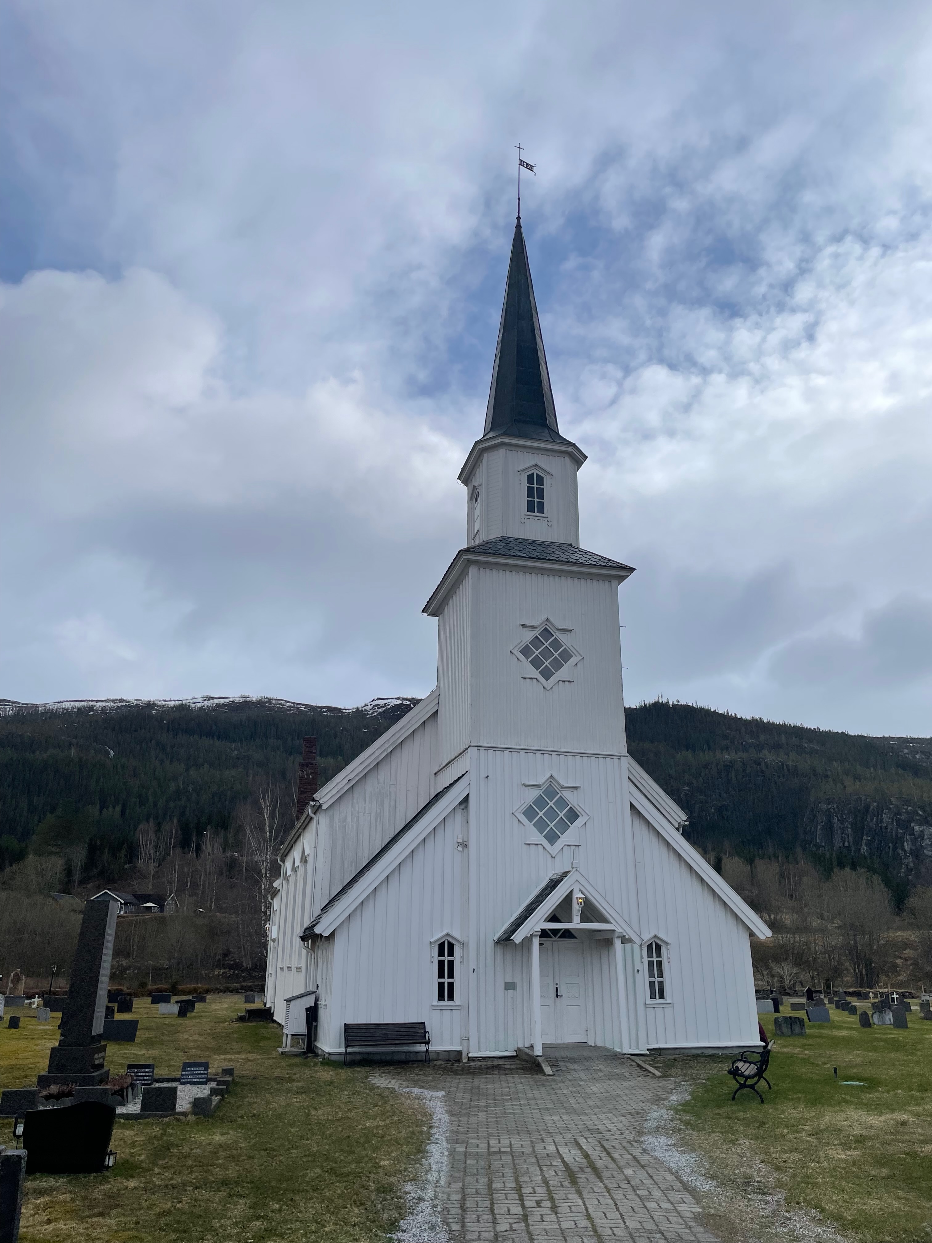 Grane Church in Grane near Trofors, photographed from the front with its white wooden entrance, tall steeple, cemetery, benches, cloudy sky, and mountain landscape behind it.