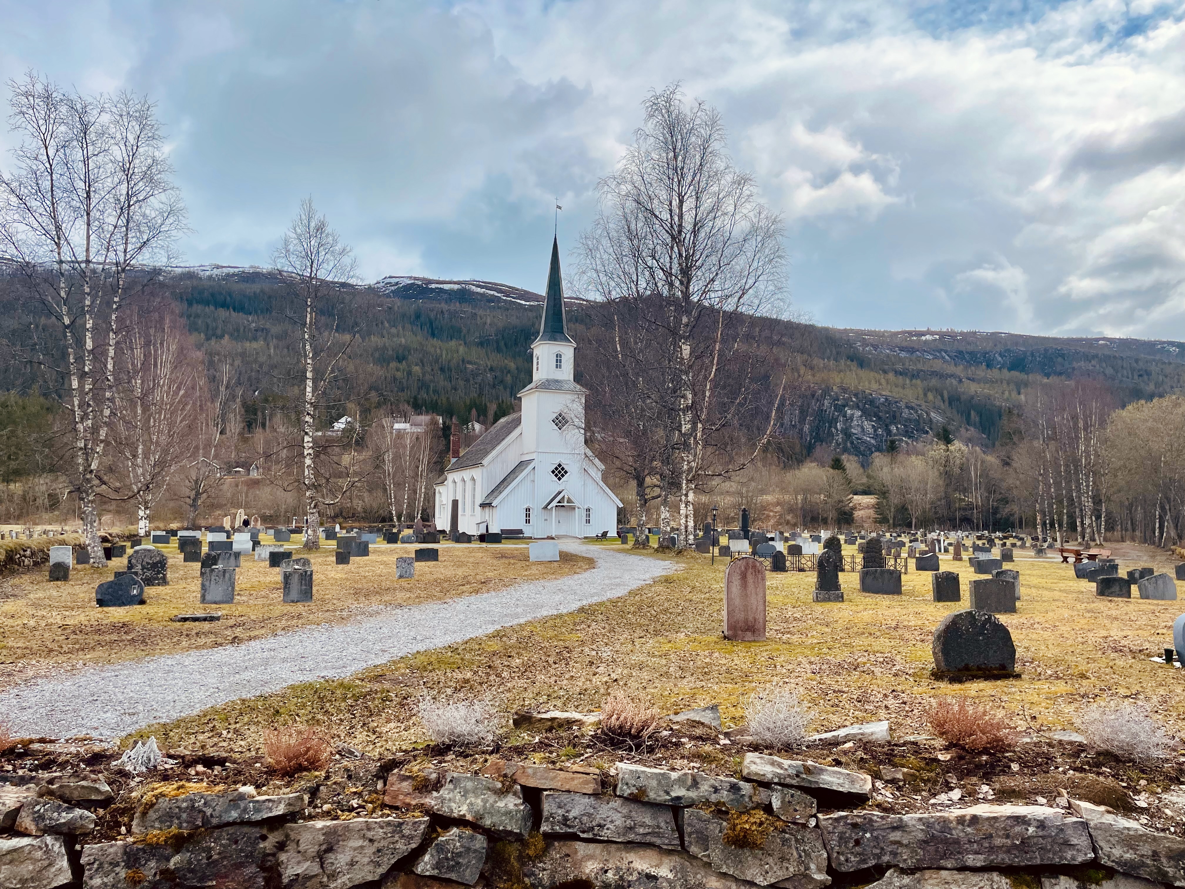 Grane Church in Grane near Trofors, a wide landscape view of the white wooden church, cemetery, gravel path, birch trees, stone wall, cloudy sky, and forested mountains in Northern Norway.