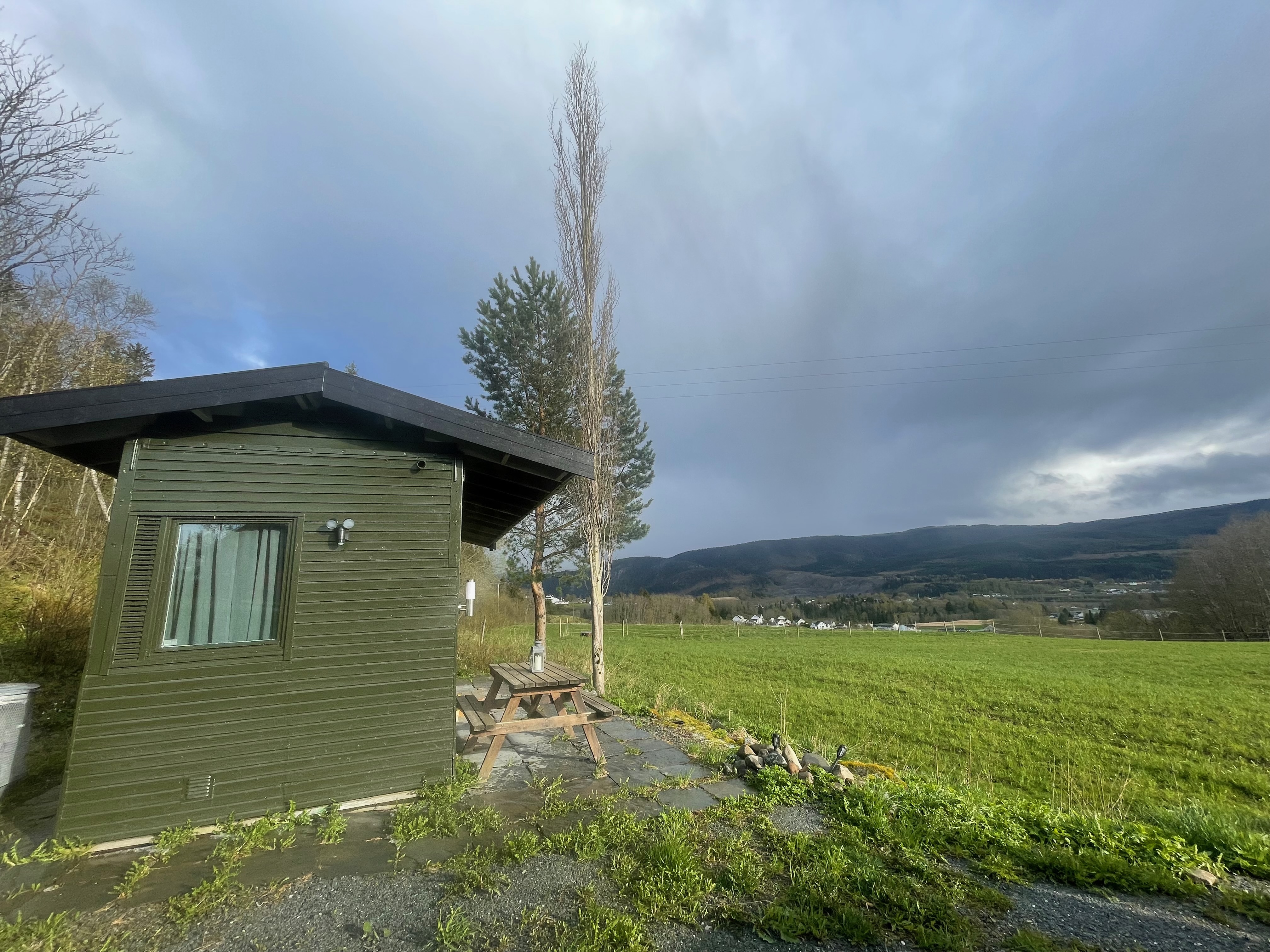 Airbnb in Stjordal Norway, green wooden cabin beside open farmland with a picnic table, cloudy sky, trees, and wide countryside views over Stjørdal.