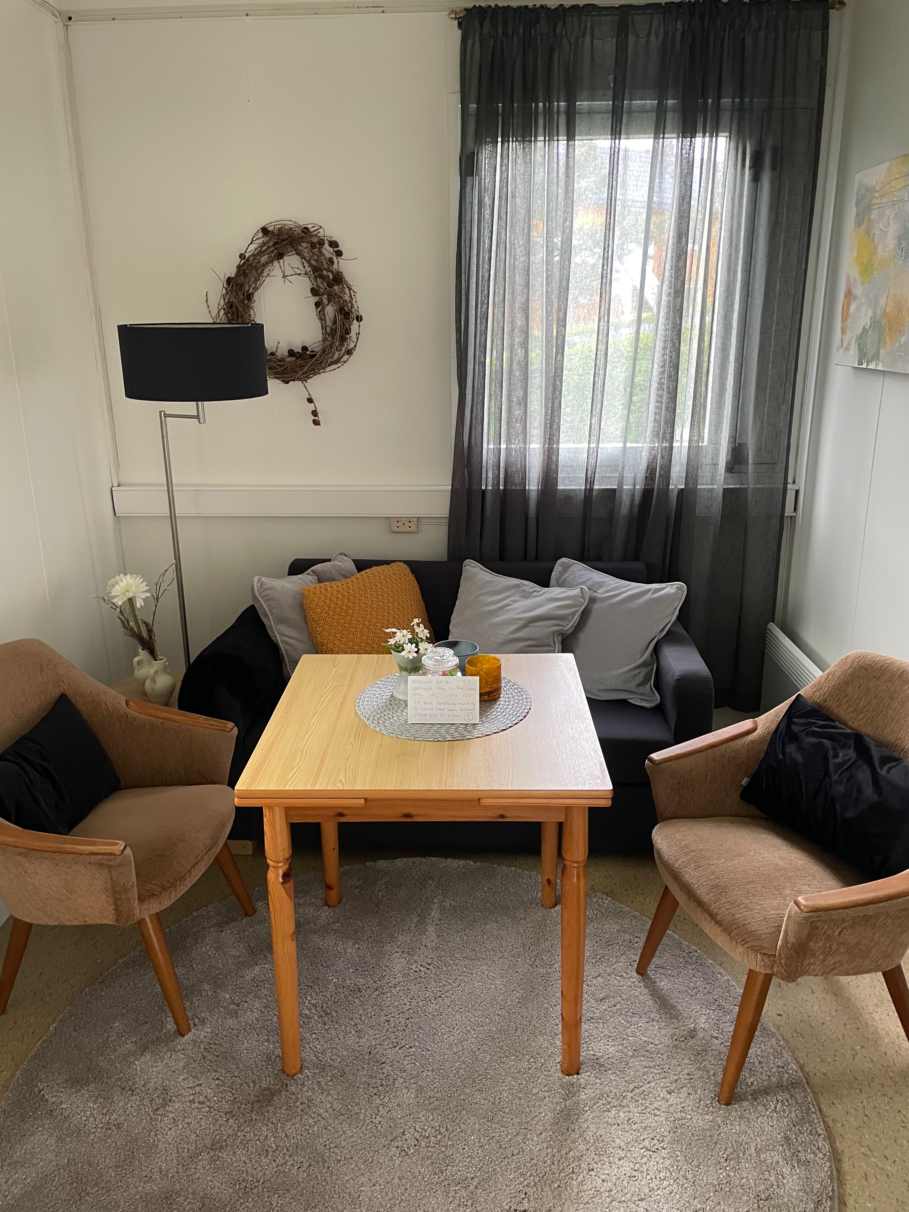 Airbnb in Stjordal Norway, cozy small living room with a wooden table, sofa, two chairs, black curtains, wall decor, and a simple countryside cabin atmosphere.