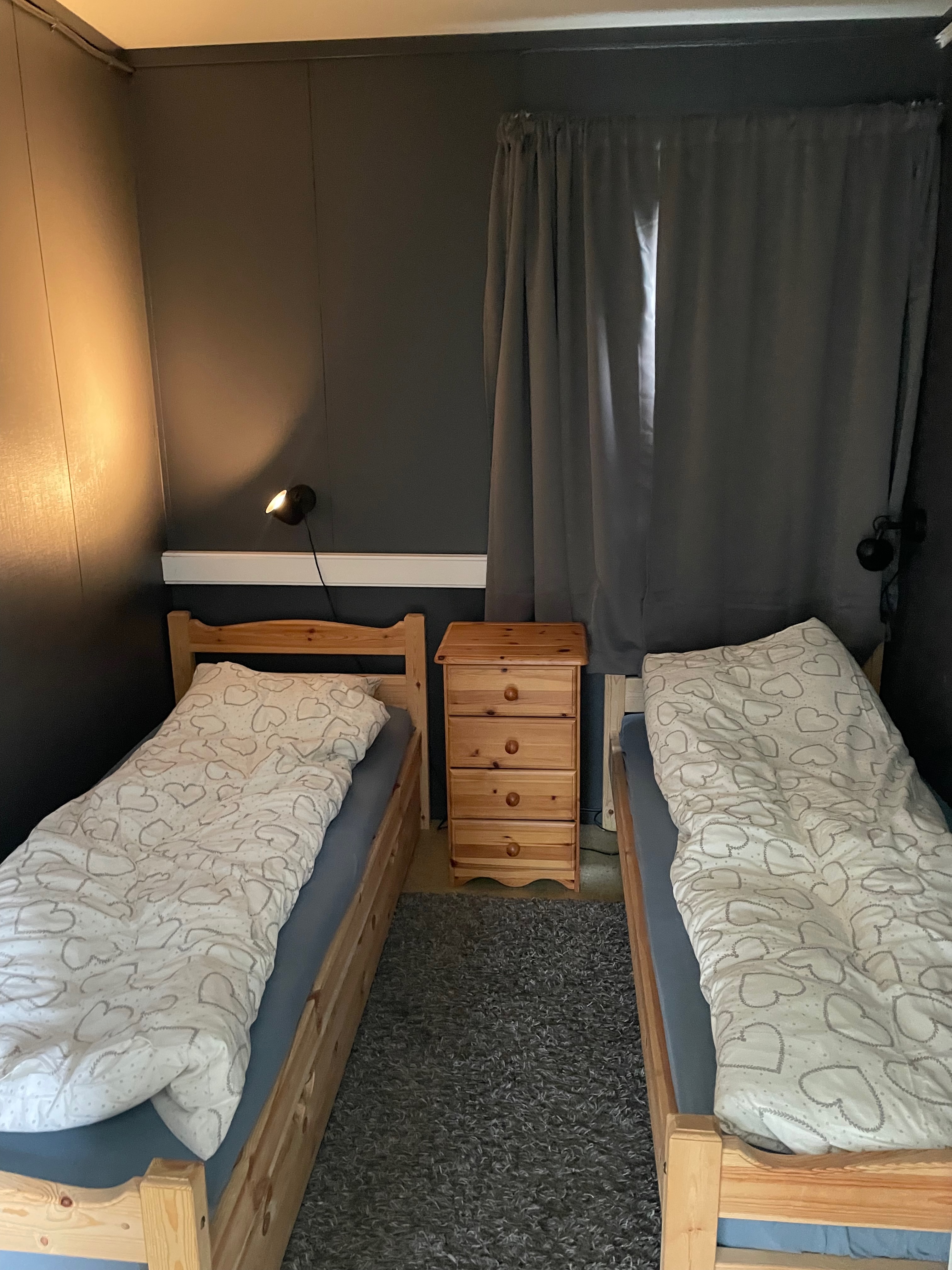 Airbnb in Stjordal Norway, small dark bedroom with two wooden single beds, heart patterned bedding, a wooden nightstand, curtains, and reading lamps.