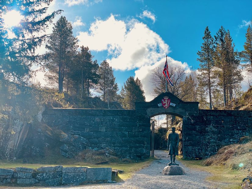 Hegra Fortress entrance gate in Norway with historic stone walls, a Norwegian flag, a soldier statue, forest scenery, and bright morning sunlight