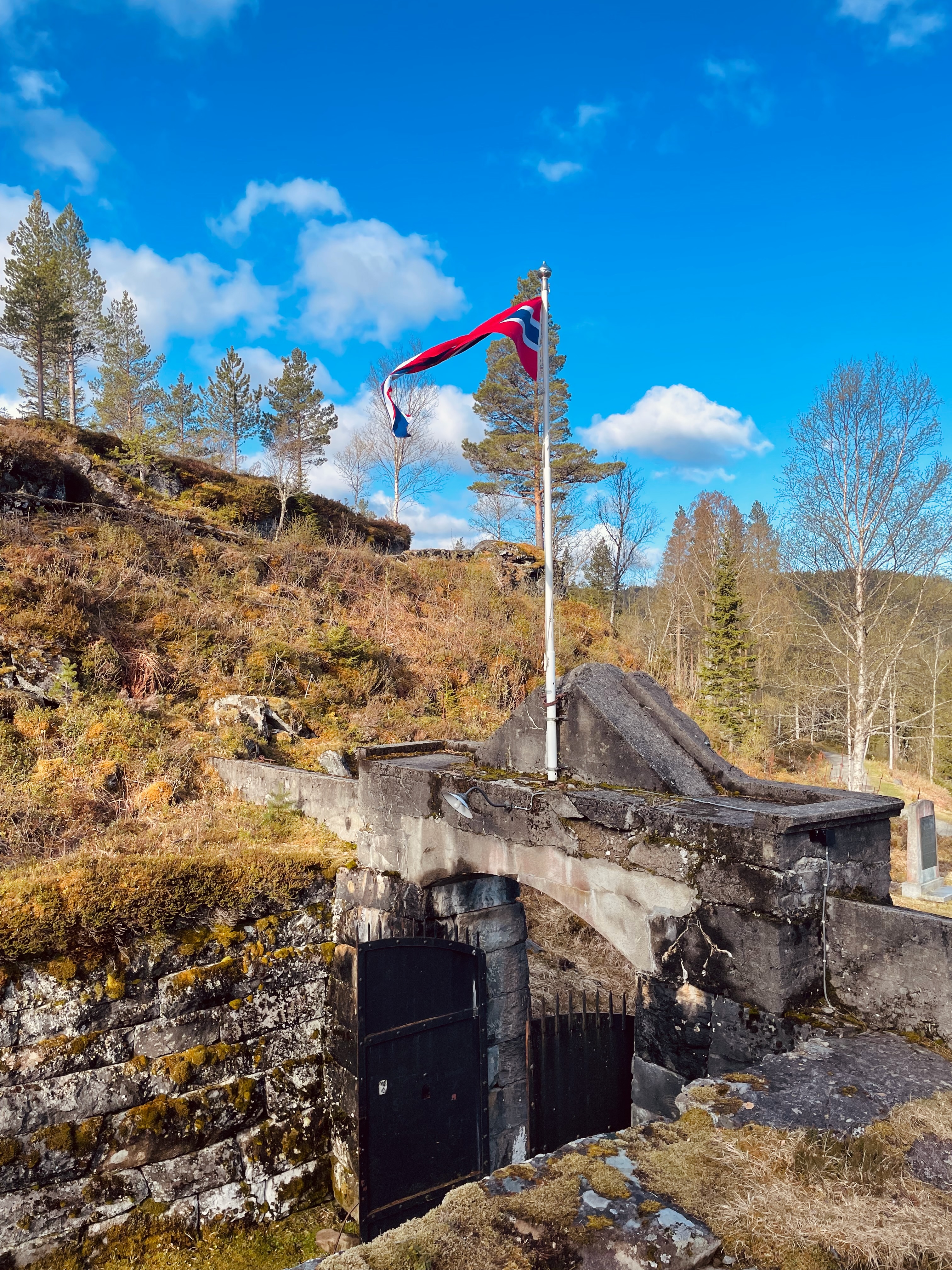Hegra Fortress entrance gate with the Norwegian flag above historic concrete walls, rocky terrain, and forest scenery in Norway.
