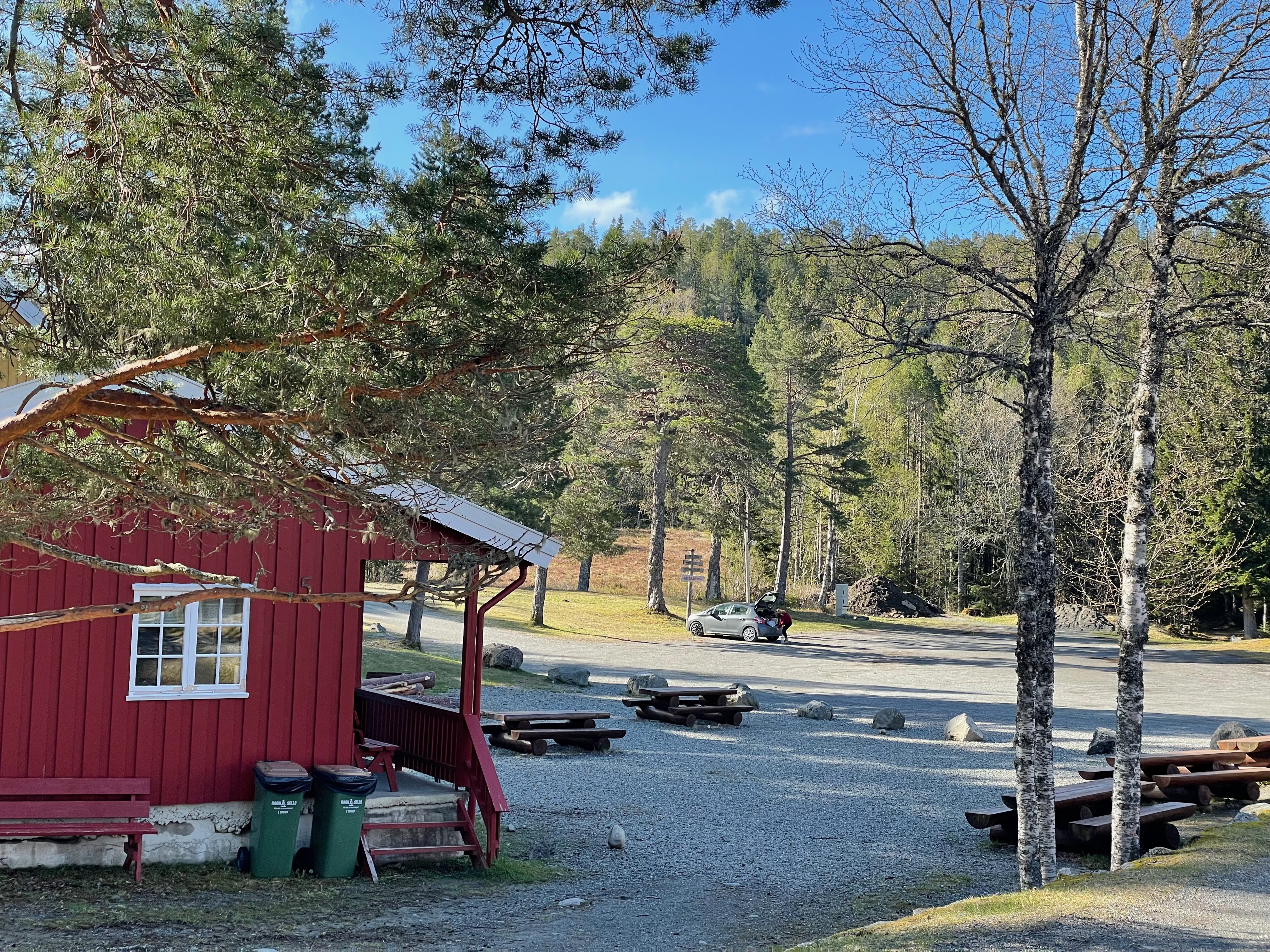 Hegra Fortress visitor area in Norway with a red wooden building, picnic benches, gravel parking space, forest scenery, and a quiet morning atmosphere