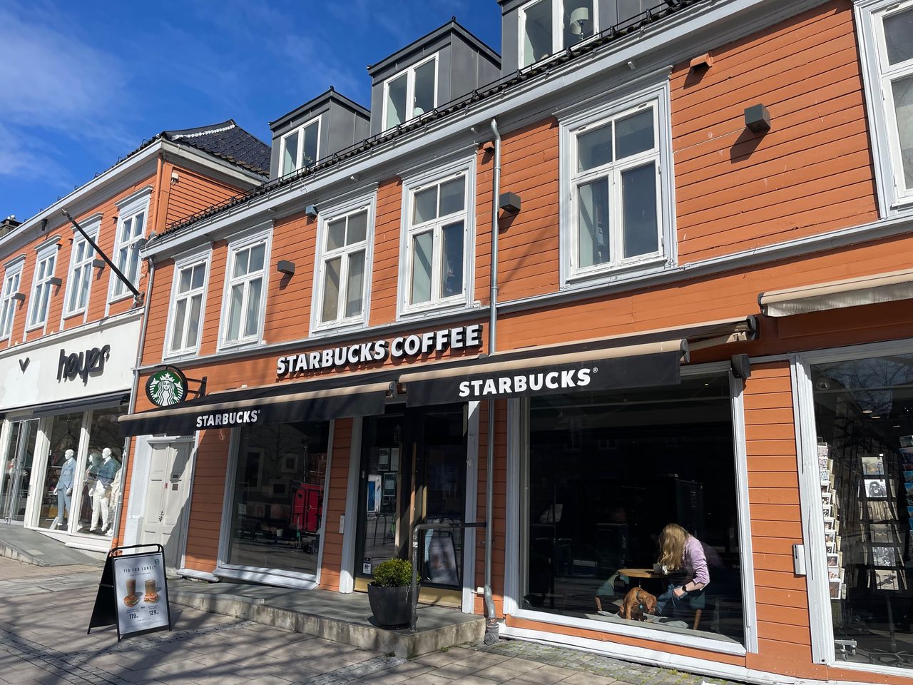 Starbucks Trondheim exterior on Kongens gate, showing the orange wooden building, black Starbucks awnings, and sunny city street.