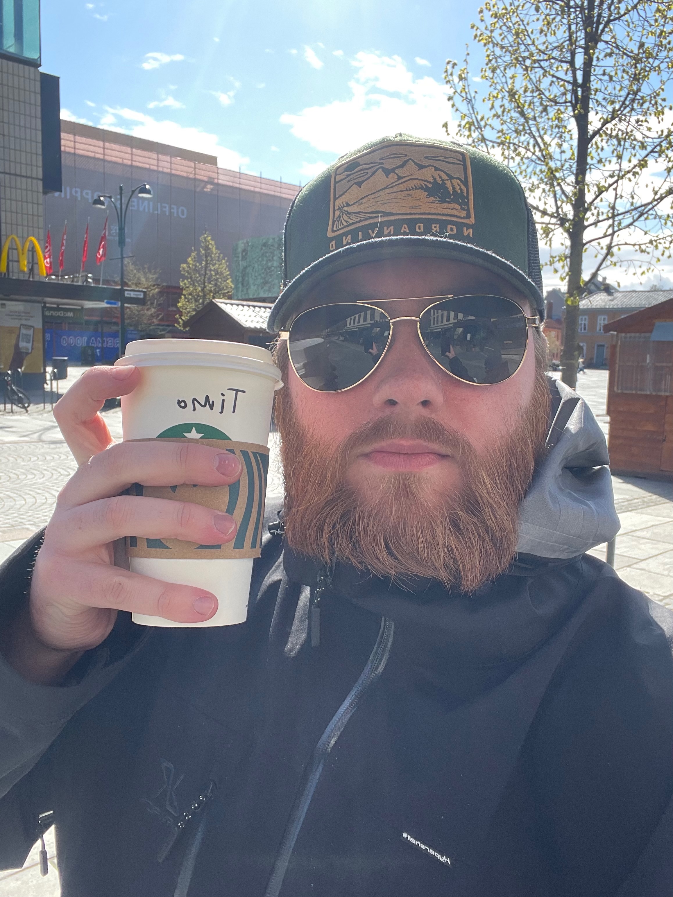Starbucks Trondheim coffee moment outside, with Timo van den Berg holding a Starbucks cup in the sunny city center of Trondheim.