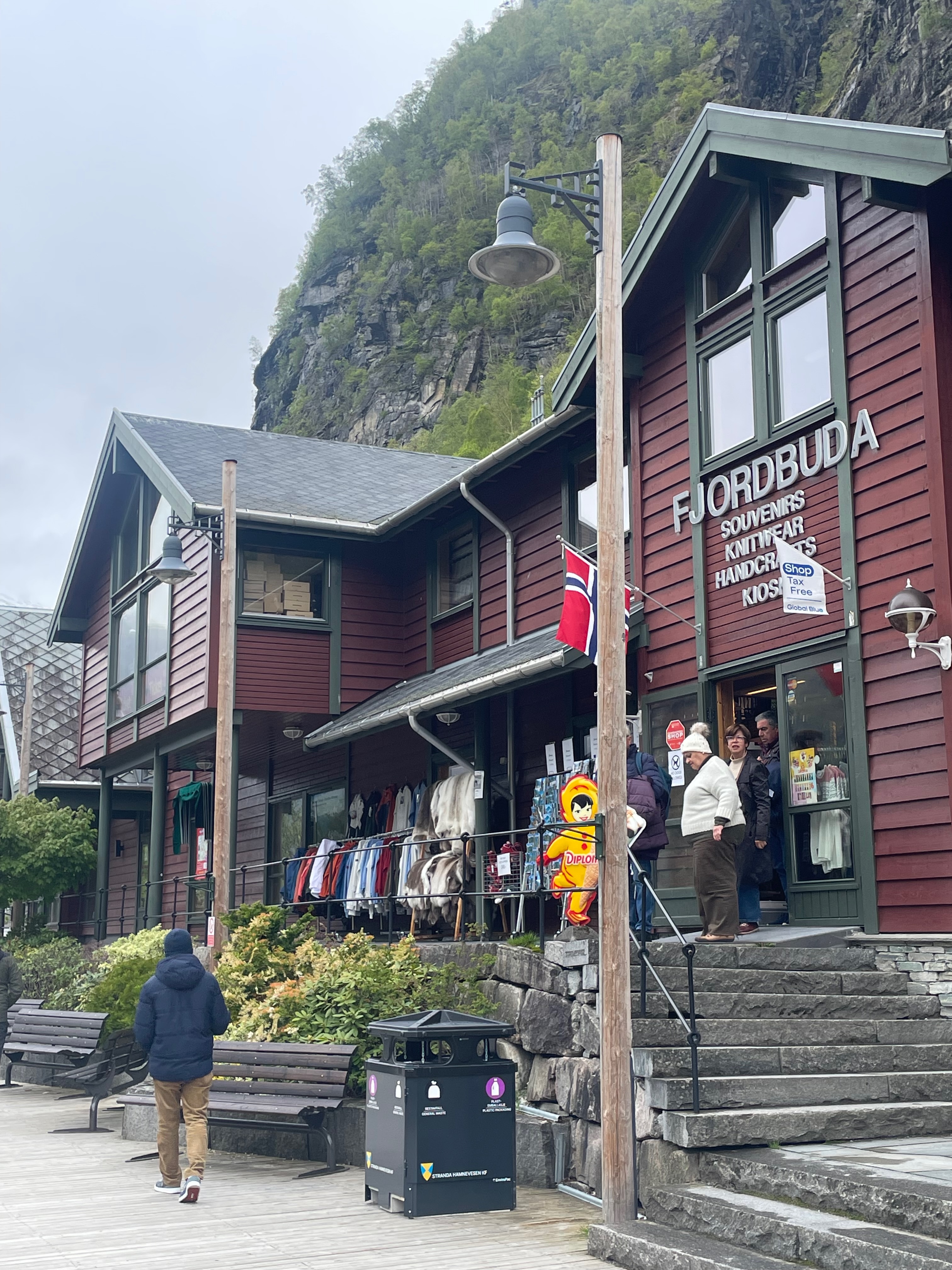 Fjordbuda Geiranger souvenir shop entrance with red wooden building, Norway flag, outdoor clothing racks, stone steps, visitors, and steep green mountain cliffs behind the store.
