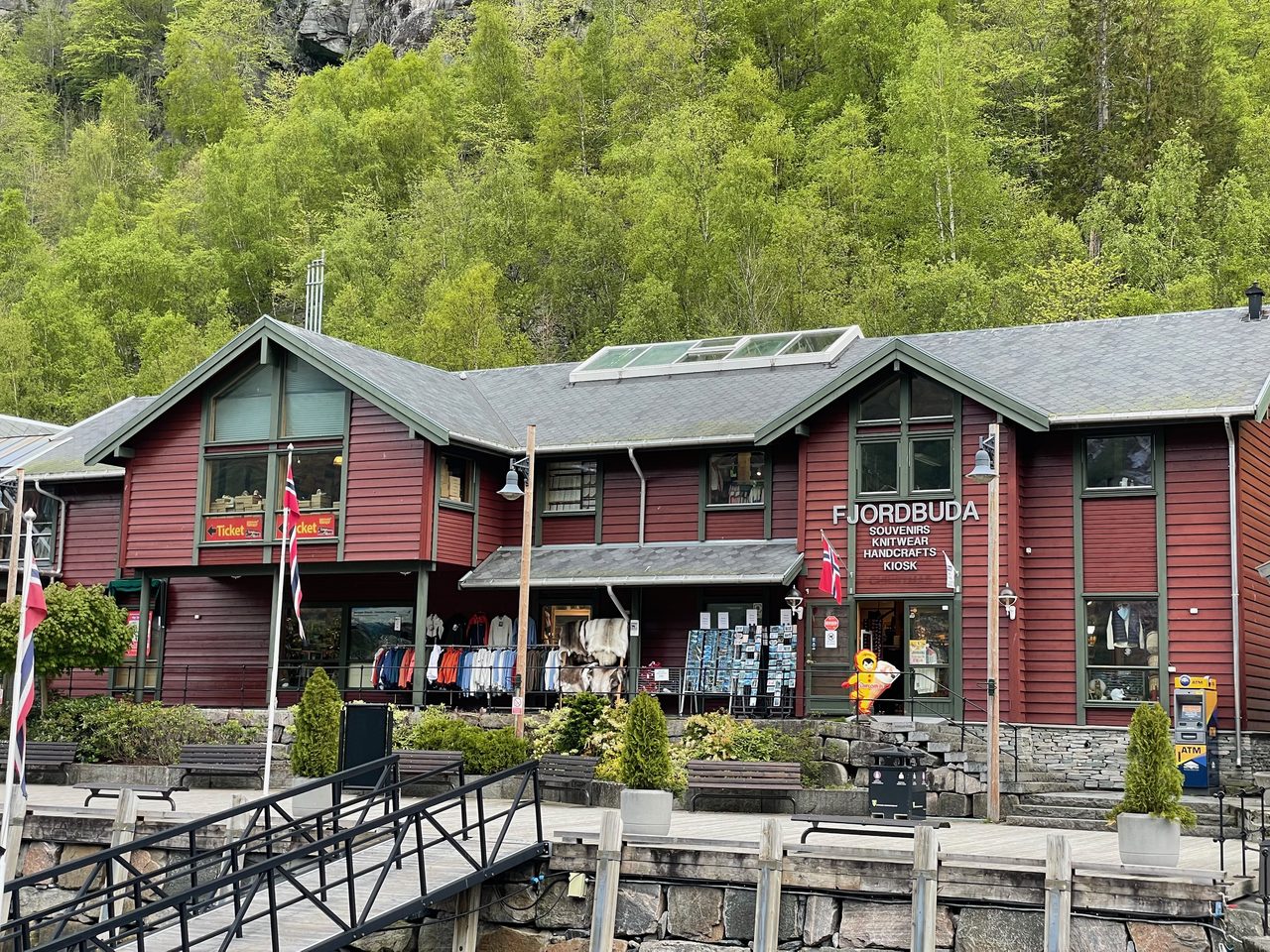 Fjordbuda Geiranger waterfront souvenir shop with red wooden facade, two floor building, benches, outdoor racks, Norway flags, and green mountain scenery in Geiranger.