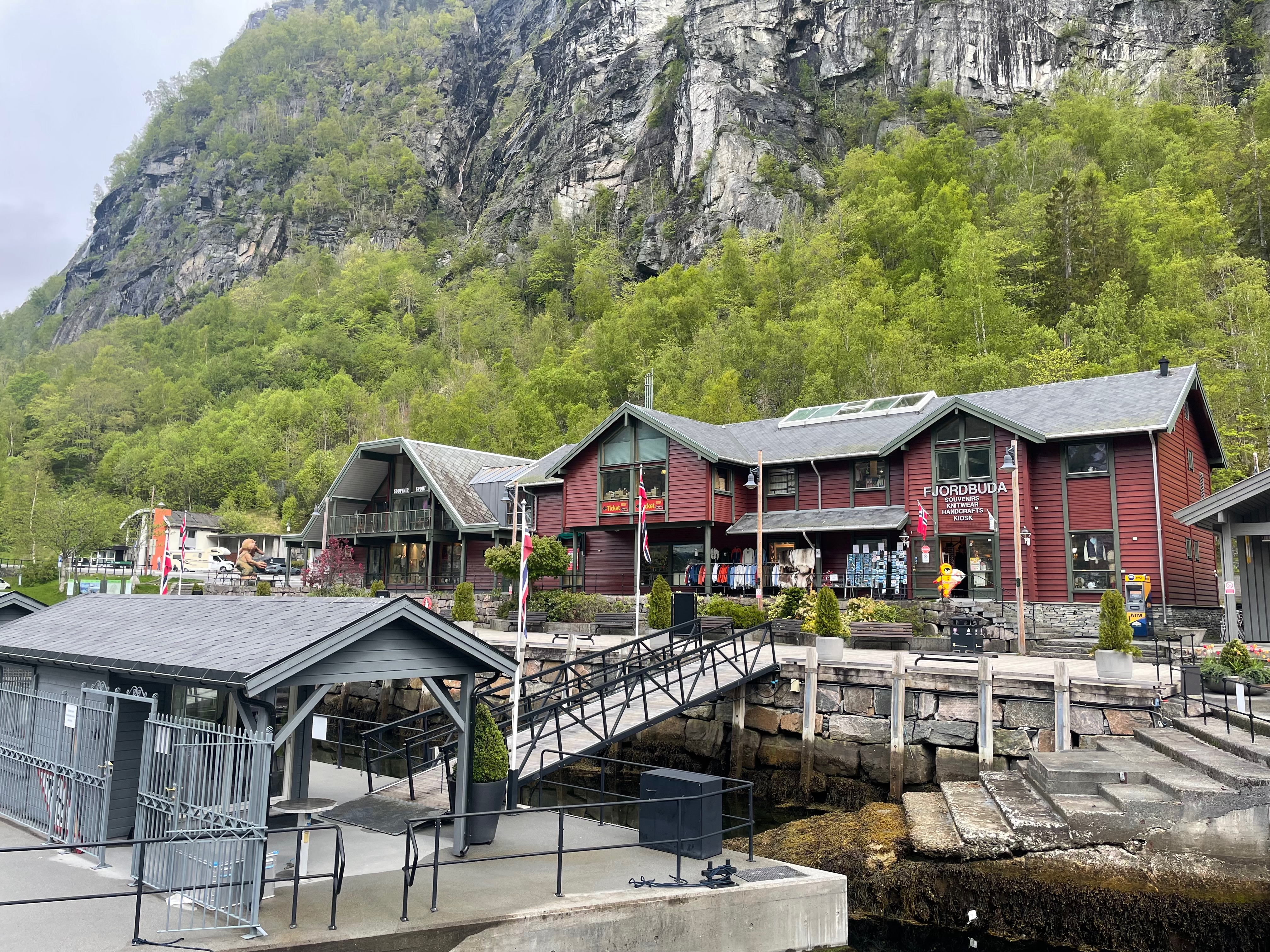 Fjordbuda Geiranger seen from the harbor area with red souvenir shop buildings, fjordside walkway, steep cliffs, green trees, and tourist facilities in Geiranger, Norway.