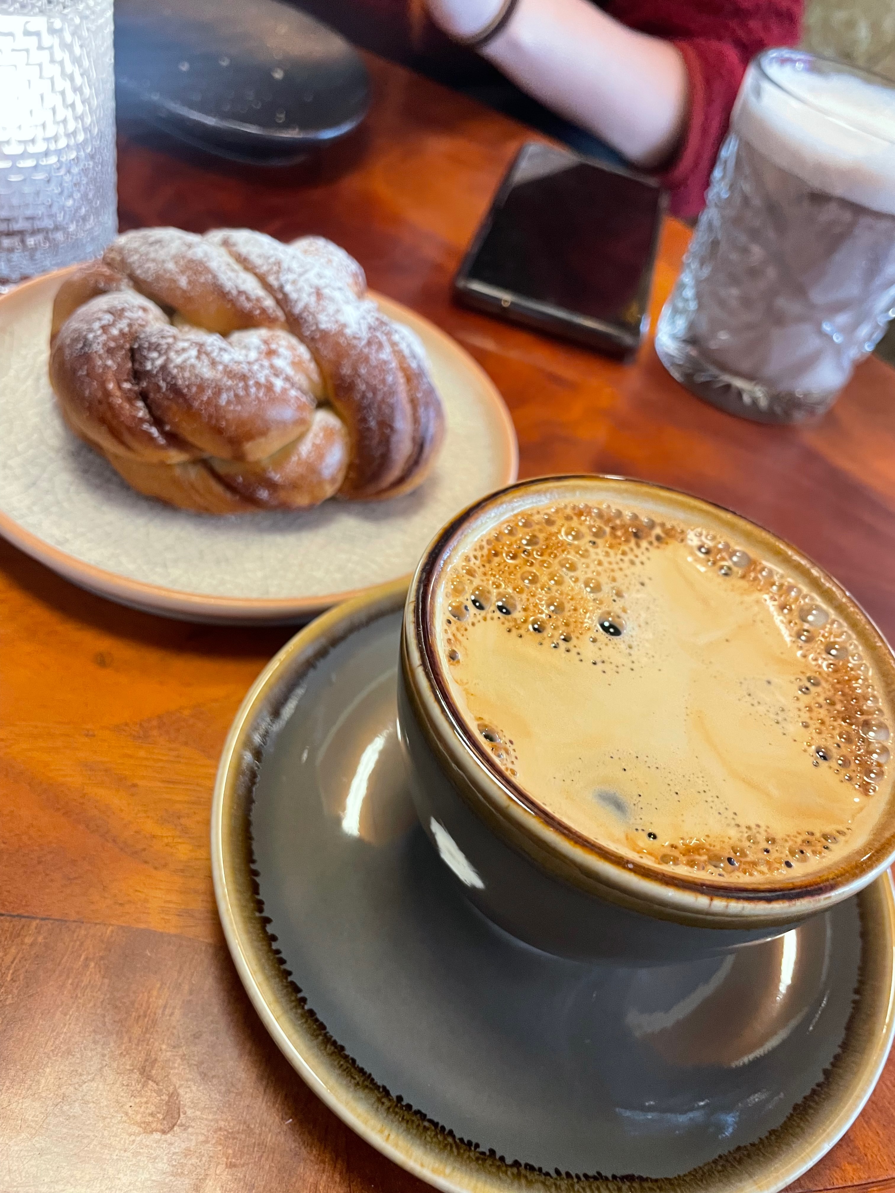 Olebuda Geiranger coffee table with a fresh Americano in a ceramic cup, a kanelknute pastry on a plate, a glass of hot chocolate, a phone, and warm wooden café table details.