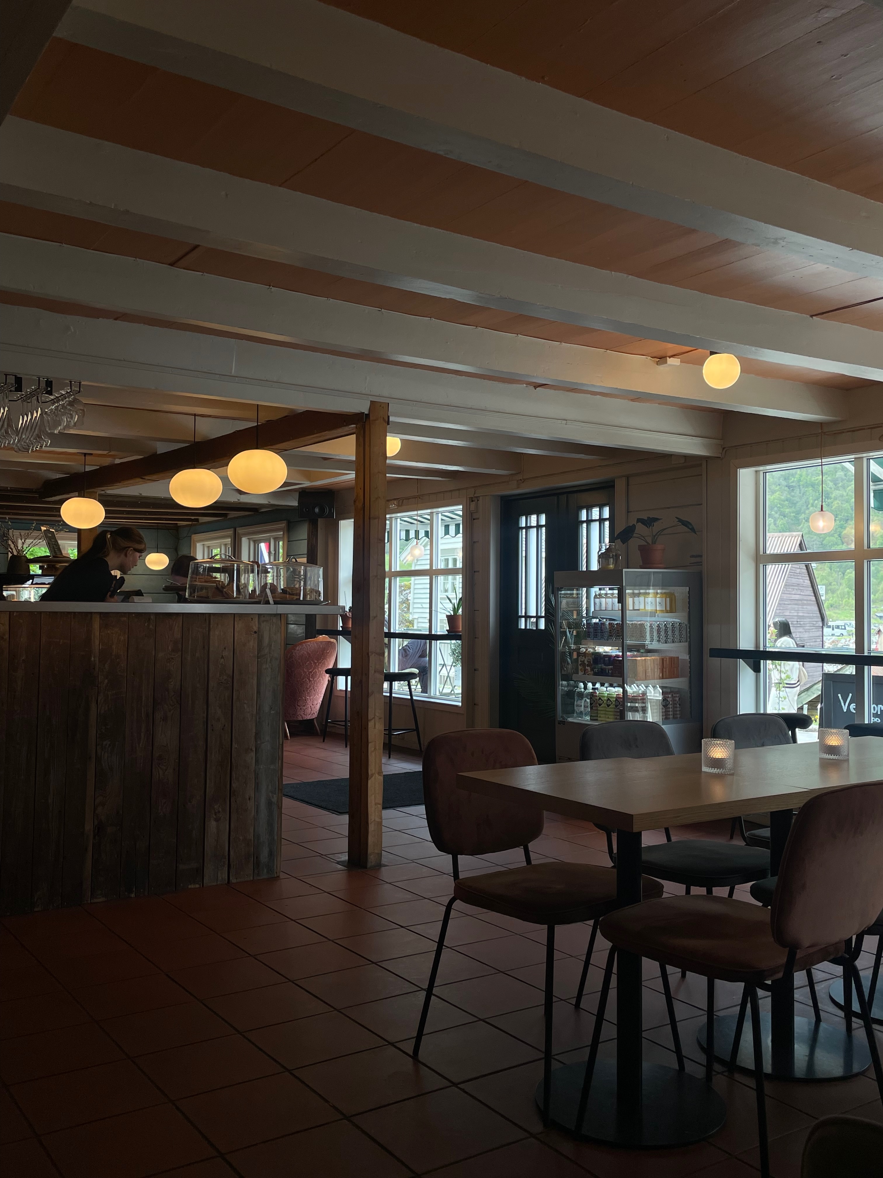 Olebuda Geiranger interior with wooden counter, warm round ceiling lights, café seating, display fridge, large windows, and a cozy fjordside café atmosphere in Norway.