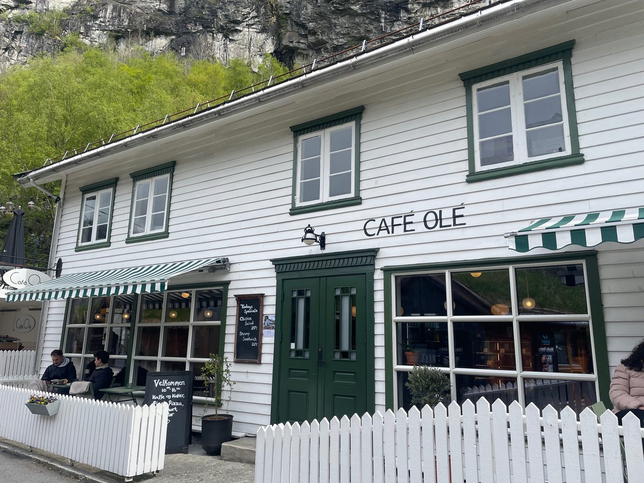 Olebuda Geiranger exterior of Café Olé with white wooden facade, green doors, striped awnings, outdoor seating, white fence, and steep rocky fjord landscape behind the building.