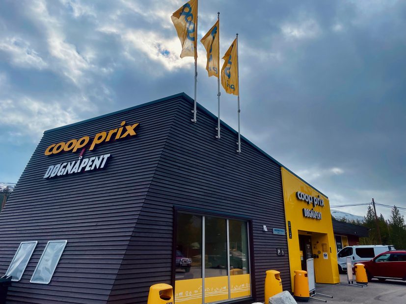 Coop Prix Bruvoll supermarket exterior in Nordberg, Norway, with yellow Coop Prix signage, Døgnåpent lettering, three yellow Coop flags, yellow entrance, parking area, bins, cars, and dramatic cloudy mountain sky