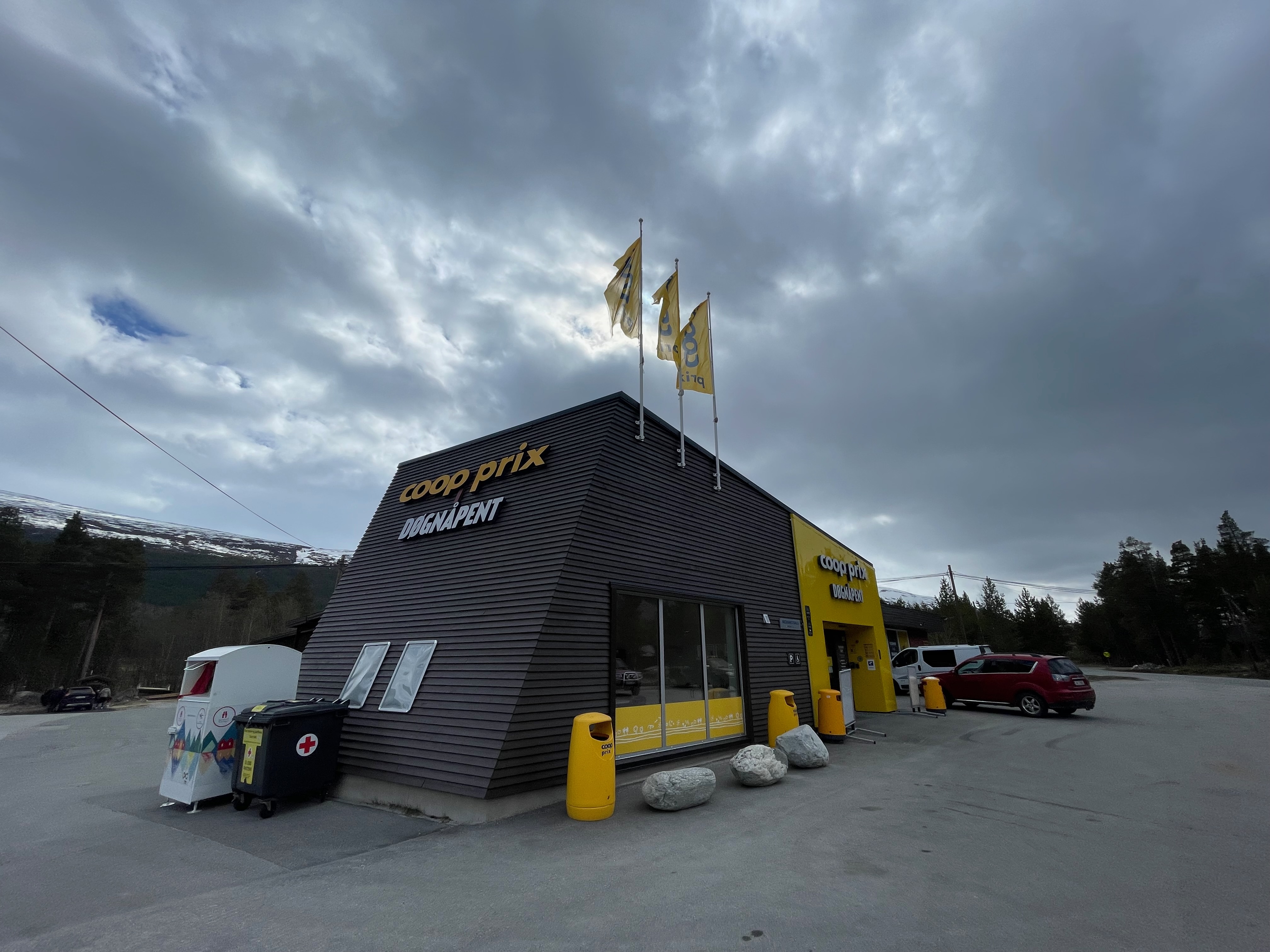 Coop Prix Bruvoll store building in Nordberg, Norway, with Coop Prix and Døgnåpent signs, yellow entrance, Coop branded trash bins, donation containers, parked cars, forest, mountains, and dark cloudy sky