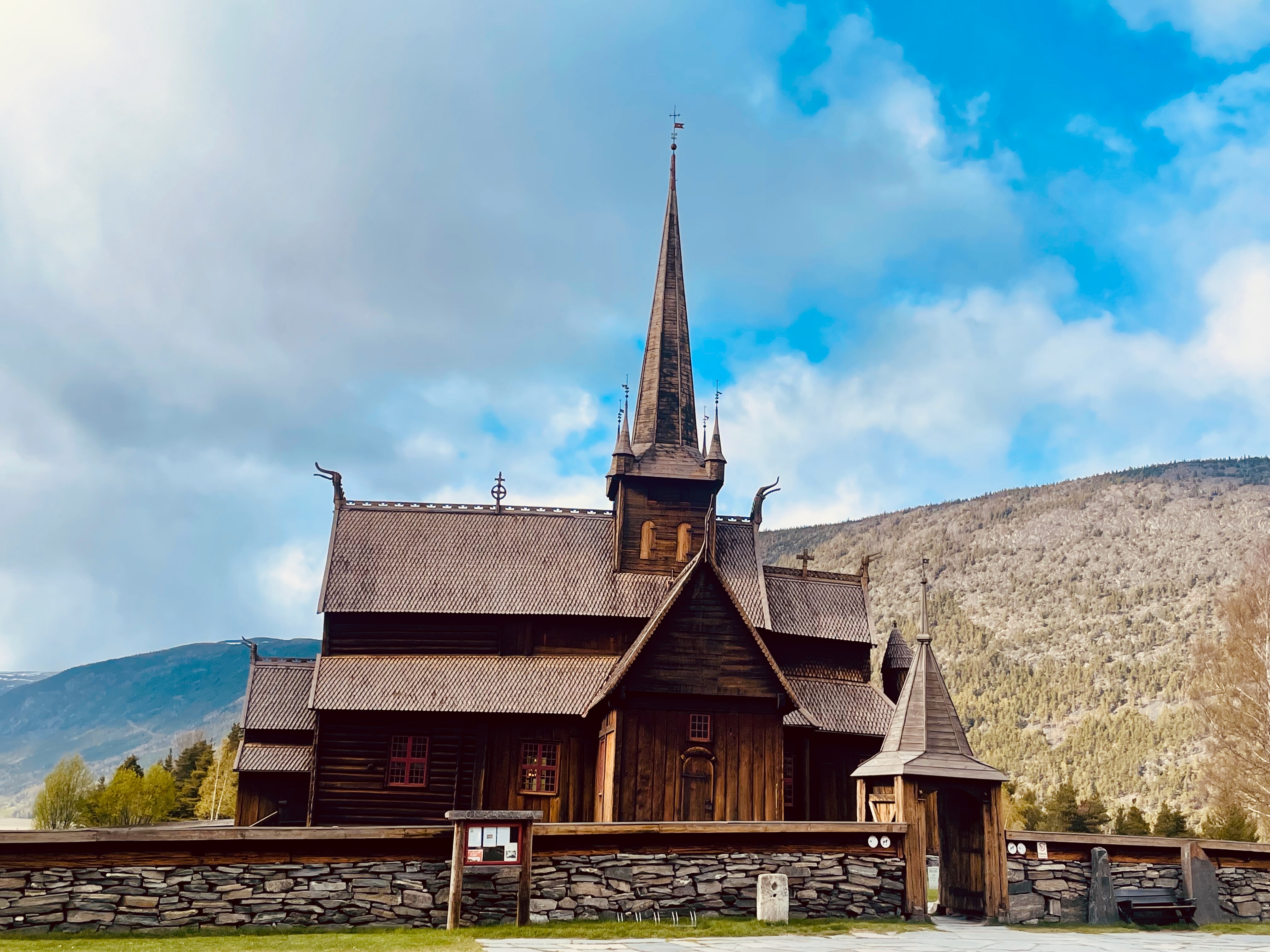 Lom Stave Church in Lom, Norway, seen from outside the stone churchyard wall with dark wooden stave architecture, tall central spire, carved roof details, wooden gatehouse, mountain landscape and dramatic blue cloudy sky.
