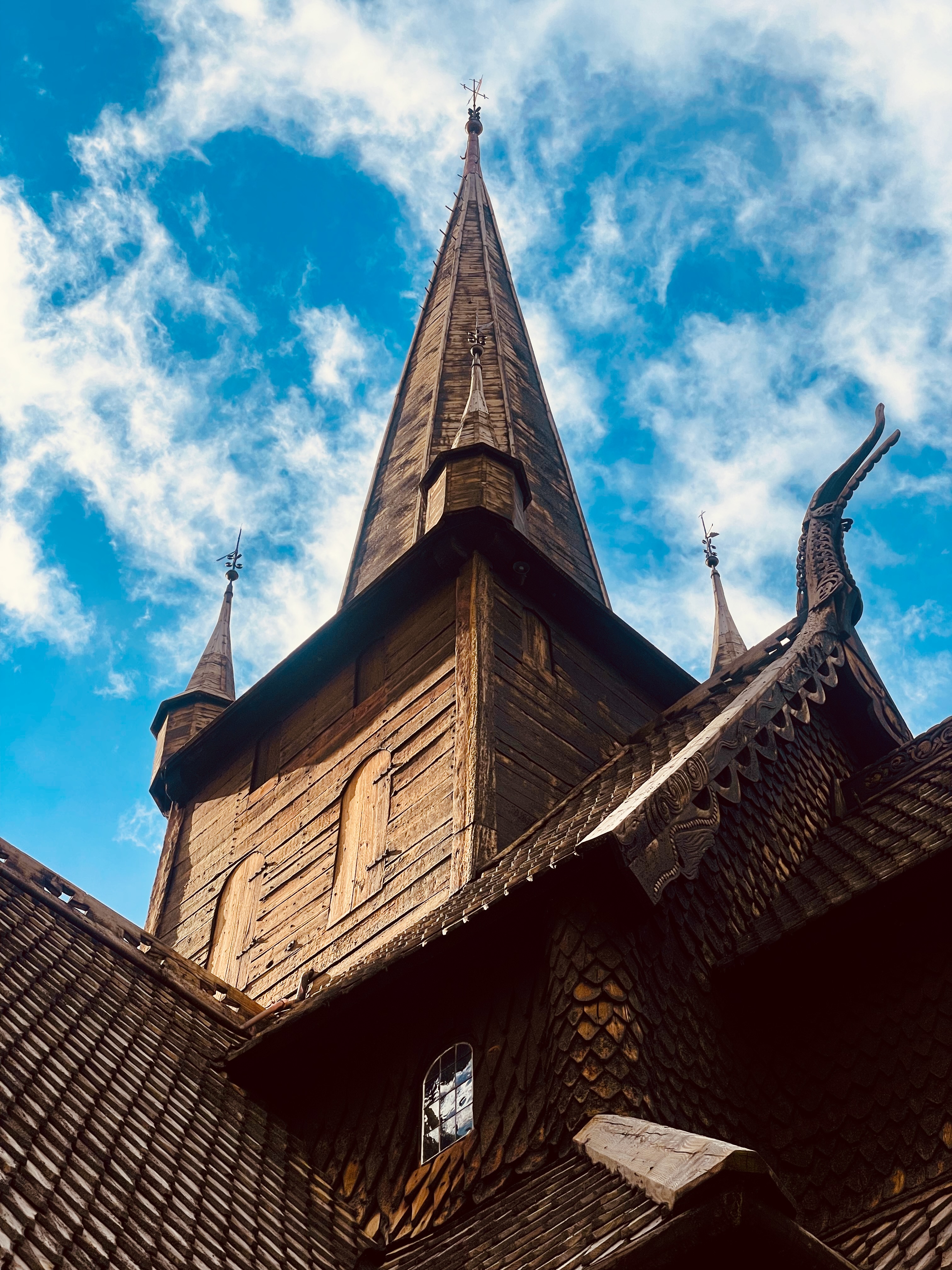 Lom Stave Church close up showing detailed medieval woodwork, dark timber roof shingles, carved dragon head decorations, wooden spires, weathered plank walls and dramatic blue cloudy sky above the church.