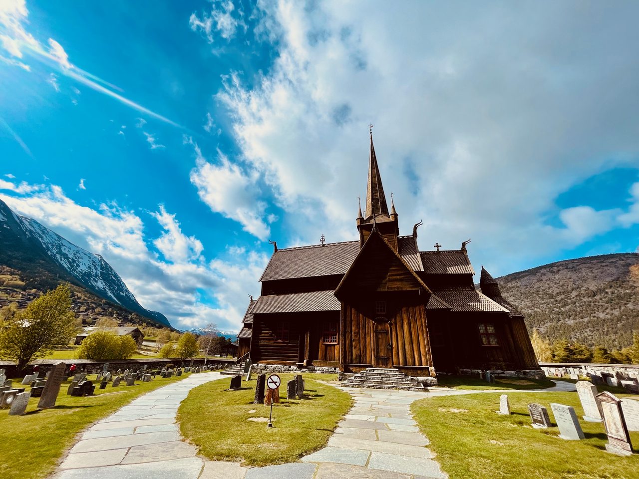 Lom Stave Church in Lom, Norway, with dark wooden stave architecture, carved roof details, a tall central spire, surrounding gravestones, mountain scenery and a dramatic blue cloudy sky.