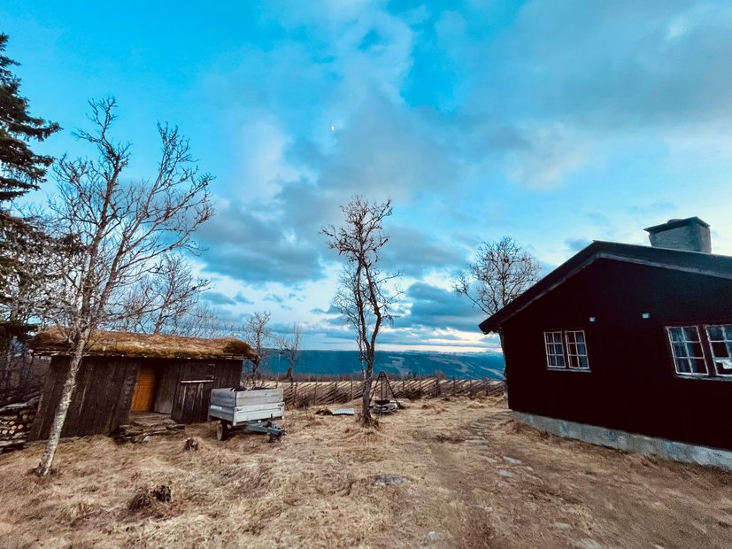 Per’s stuggu Sør-Fron mountain cabin in Norway with a dark red wooden cabin, small turf roof shed, fire pit, trailer, bare trees, mountain views and cloudy blue evening sky.