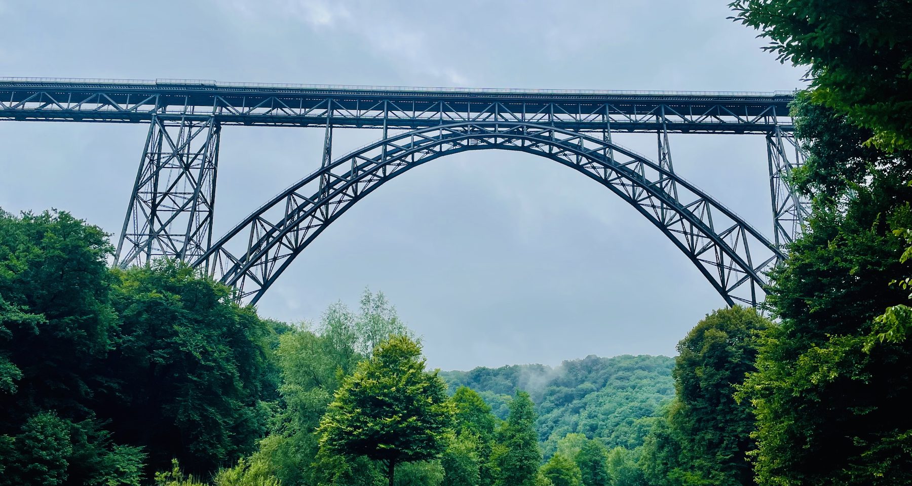 Müngstener Brücke towering above the green Brückenpark Müngsten valley in Solingen, Germany, with forest views and cloudy skies.