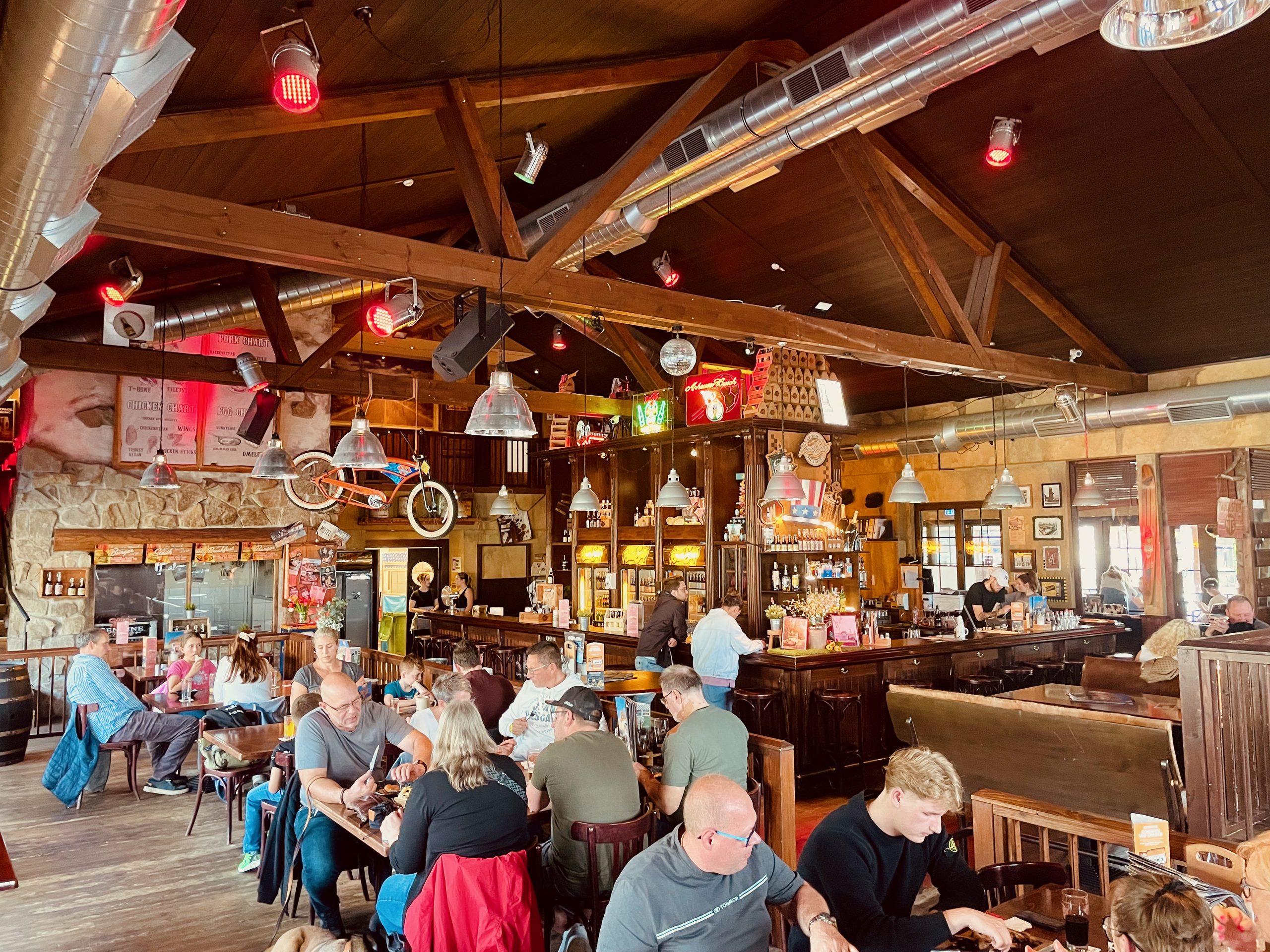 Road Stop Wuppertal interior with a busy American style restaurant, wooden beams, bar area, hanging lights, neon signs, and guests dining at tables.