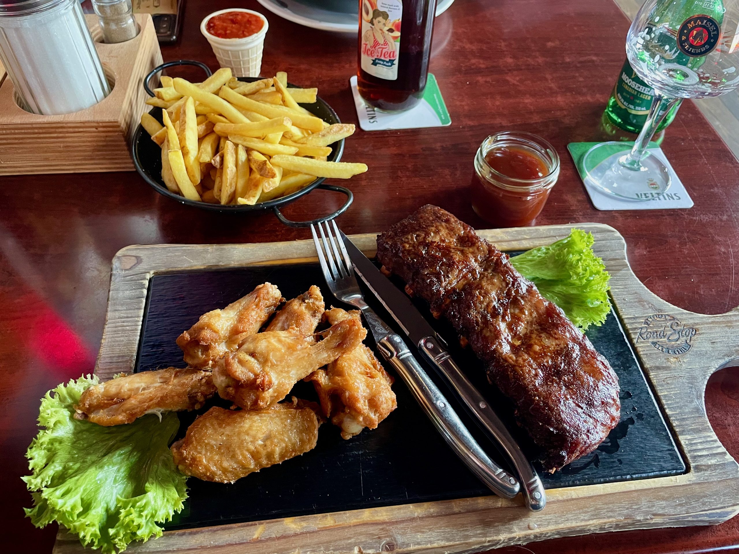 Road Stop Wuppertal dinner with Texas Ribs, chicken wings, fries, BBQ sauce, beer, and water served on a wooden platter inside the American style restaurant.