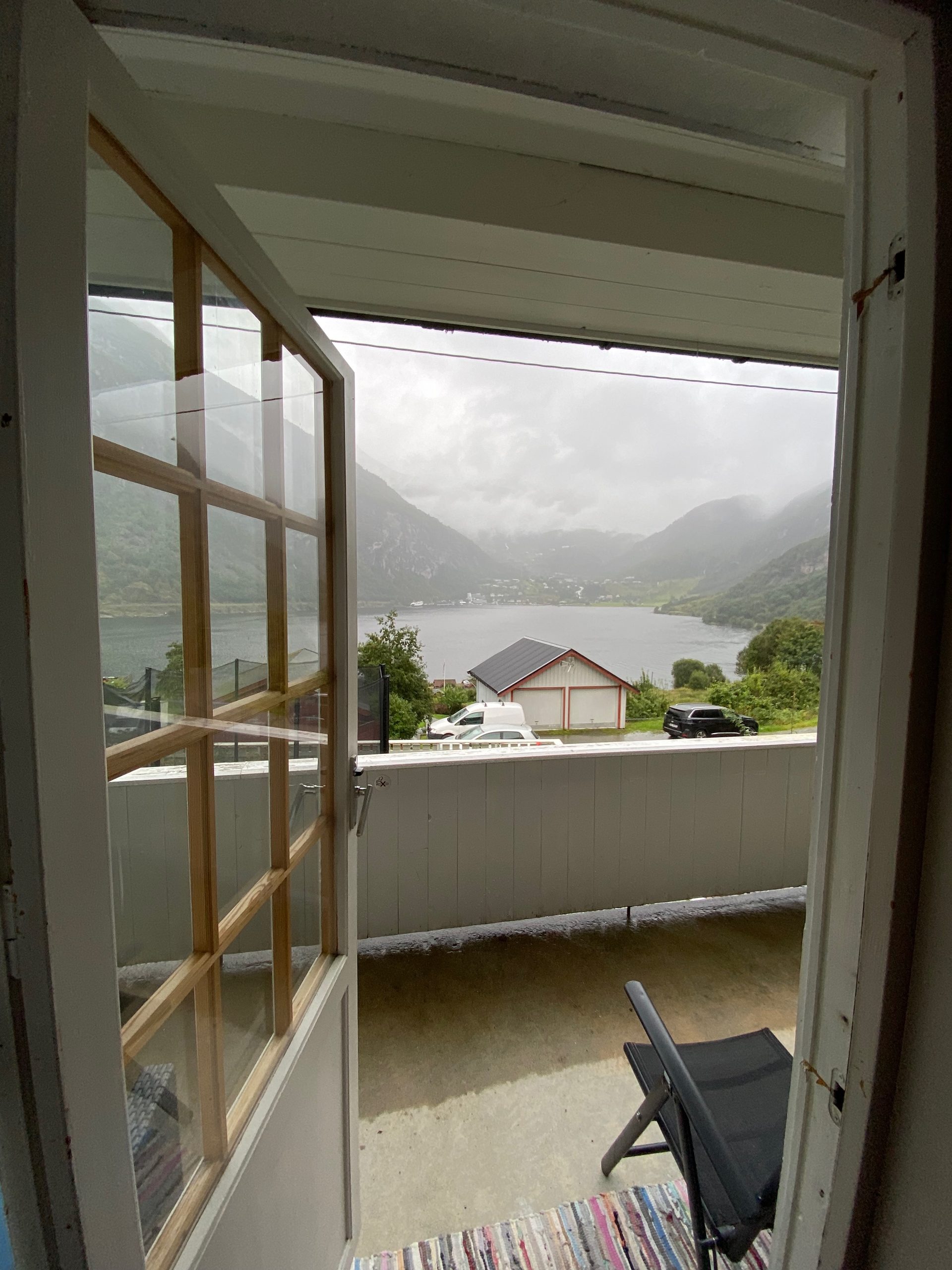 Geiranger Airbnb balcony view from the apartment doorway overlooking Geirangerfjord, cloudy mountains, waterfalls, and the village of Geiranger in Norway