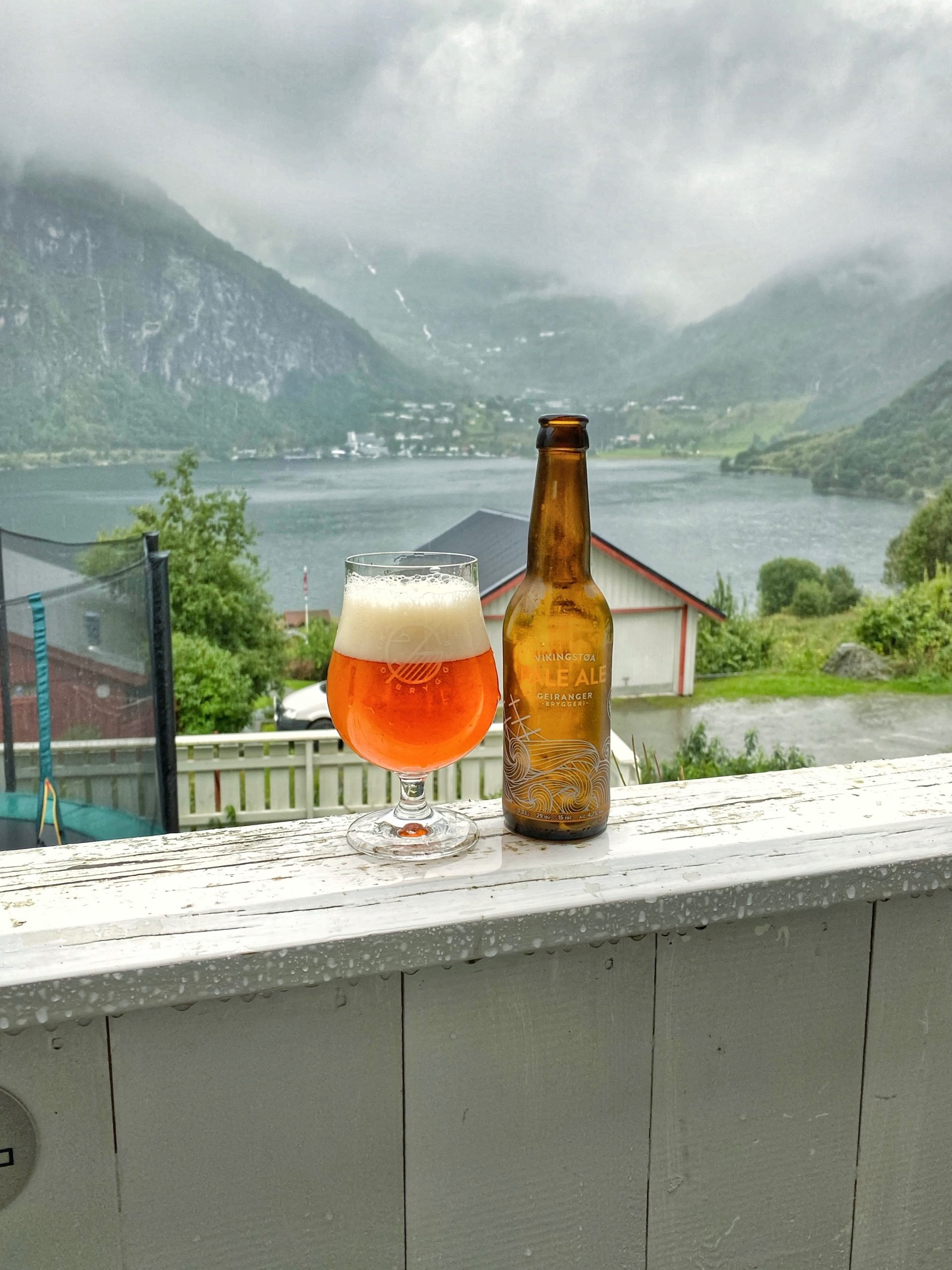 Geiranger Airbnb balcony drink with local Geiranger pale ale overlooking Geirangerfjord, misty mountains, and rainy fjord scenery