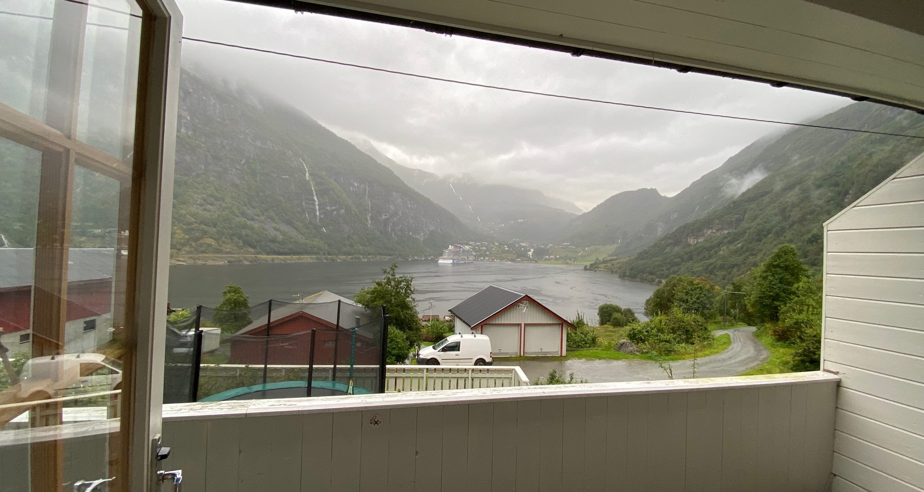 Geiranger Airbnb balcony view overlooking Geirangerfjord, mountain waterfalls, cloudy fjord weather, and the village of Geiranger in Norway