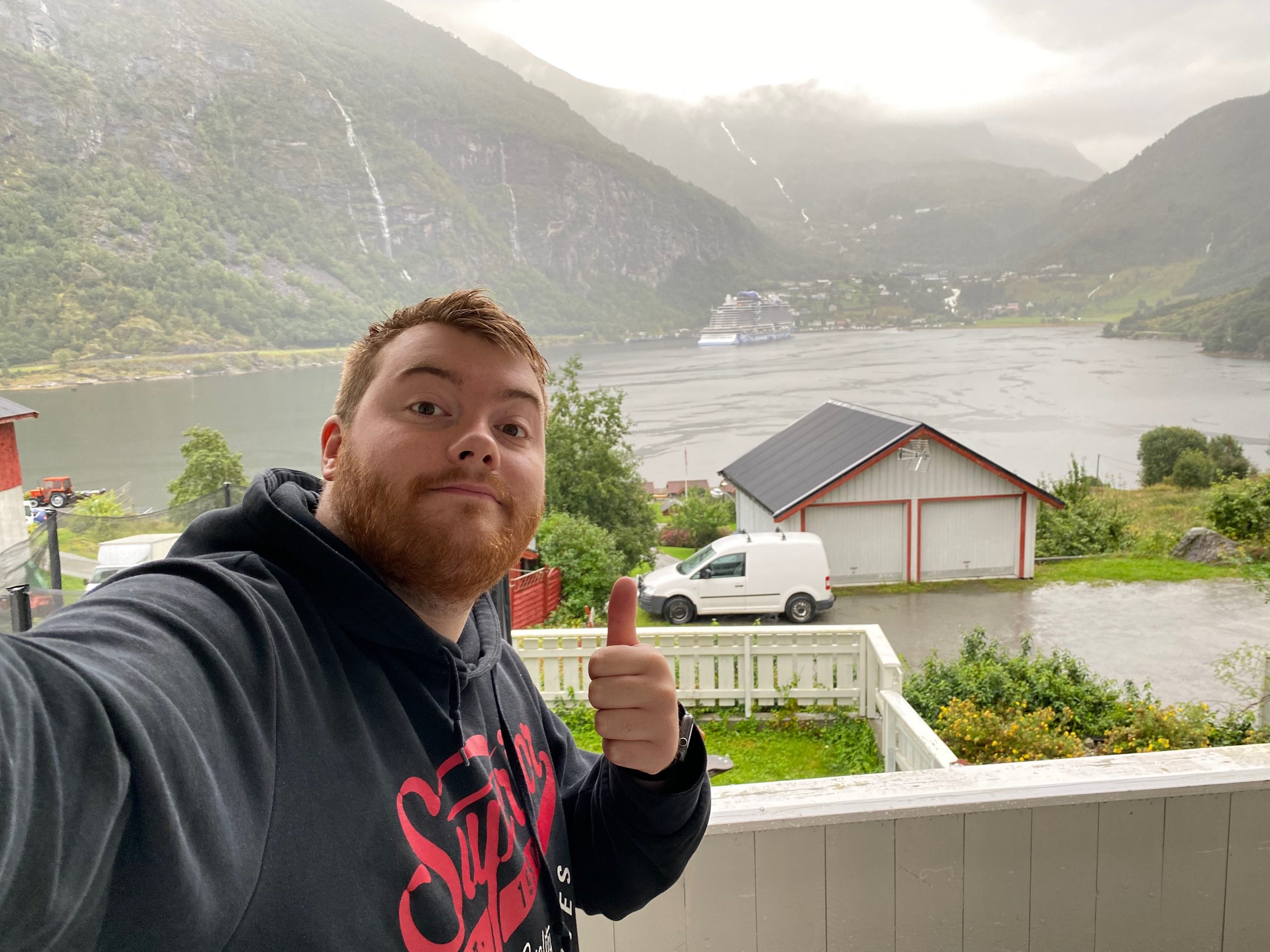 Timo van den Berg taking a selfie at a Geiranger Airbnb balcony with thumbs up, overlooking Geirangerfjord, mountain waterfalls, a cruise ship, and cloudy Norwegian fjord scenery