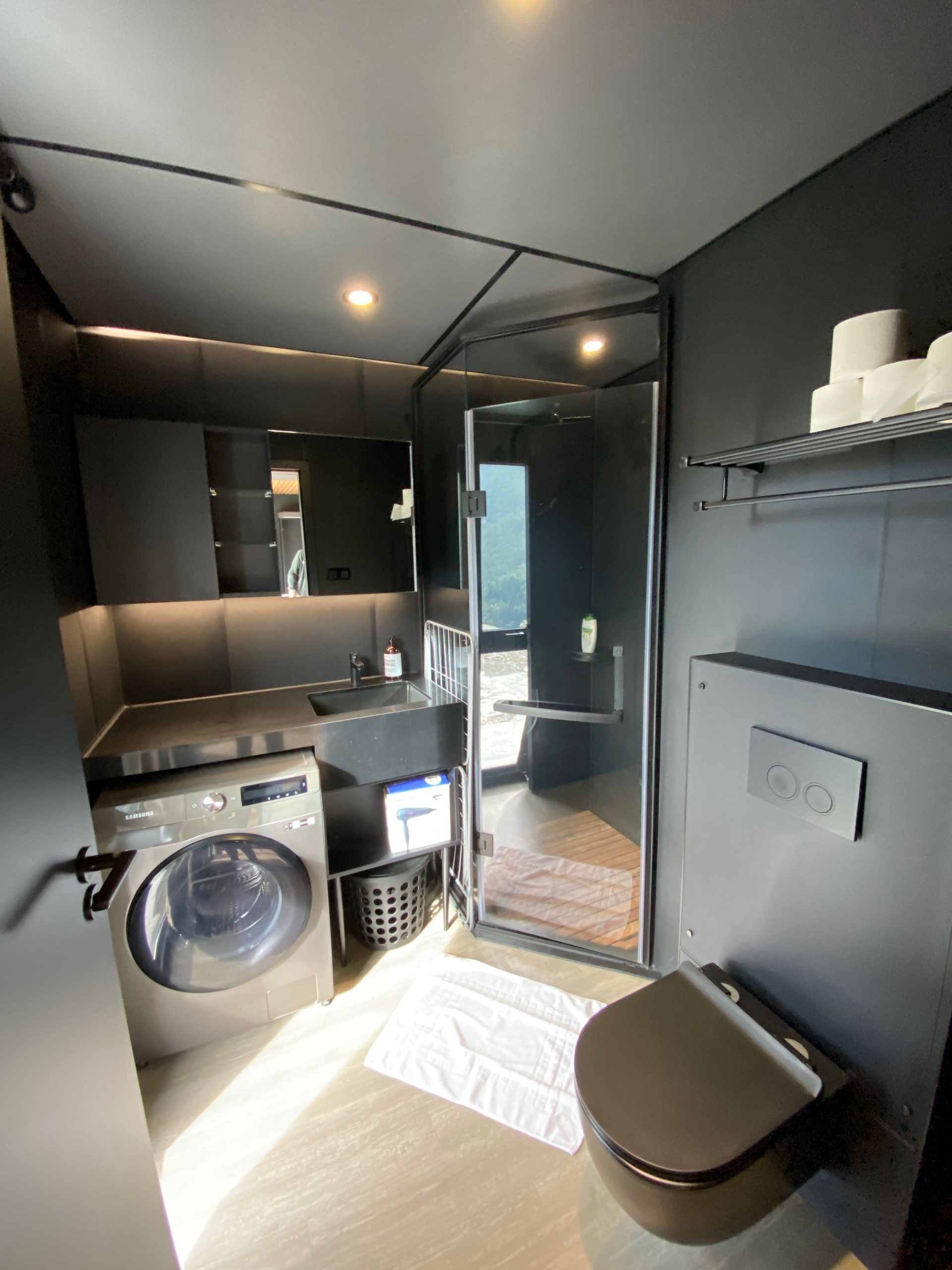 Modern dark bathroom at Sogndal Fjordpanorama cabin during a Sogndal Fjordpanorama Review, with washing machine, toilet, sink, and glass shower