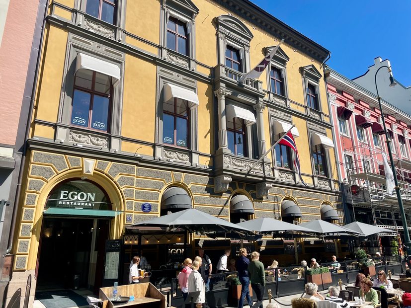 Egon Karl Johan exterior with terrace seating on Karl Johans gate in Oslo showing the yellow historic facade, Norwegian flag, umbrellas, and guests having lunch.