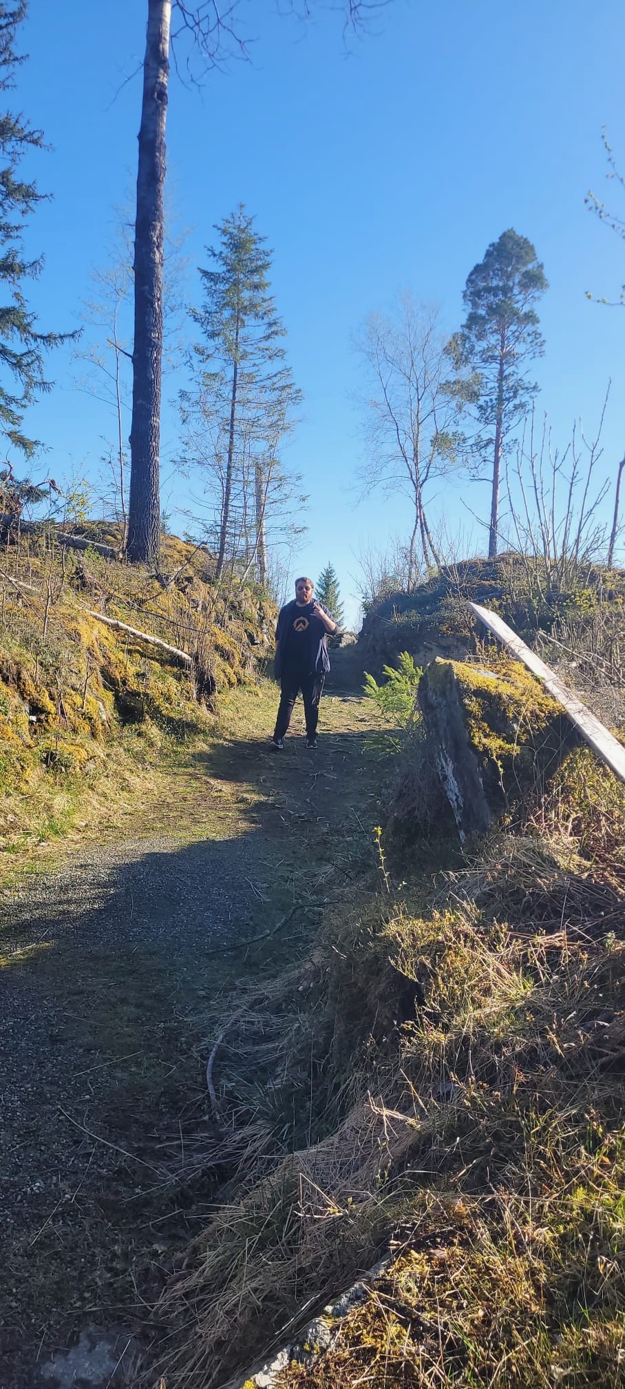 Traveler walking along a forest path at Hegra Fortress in Norway, surrounded by mossy ruins, rocky terrain, tall trees, and bright morning sunlight