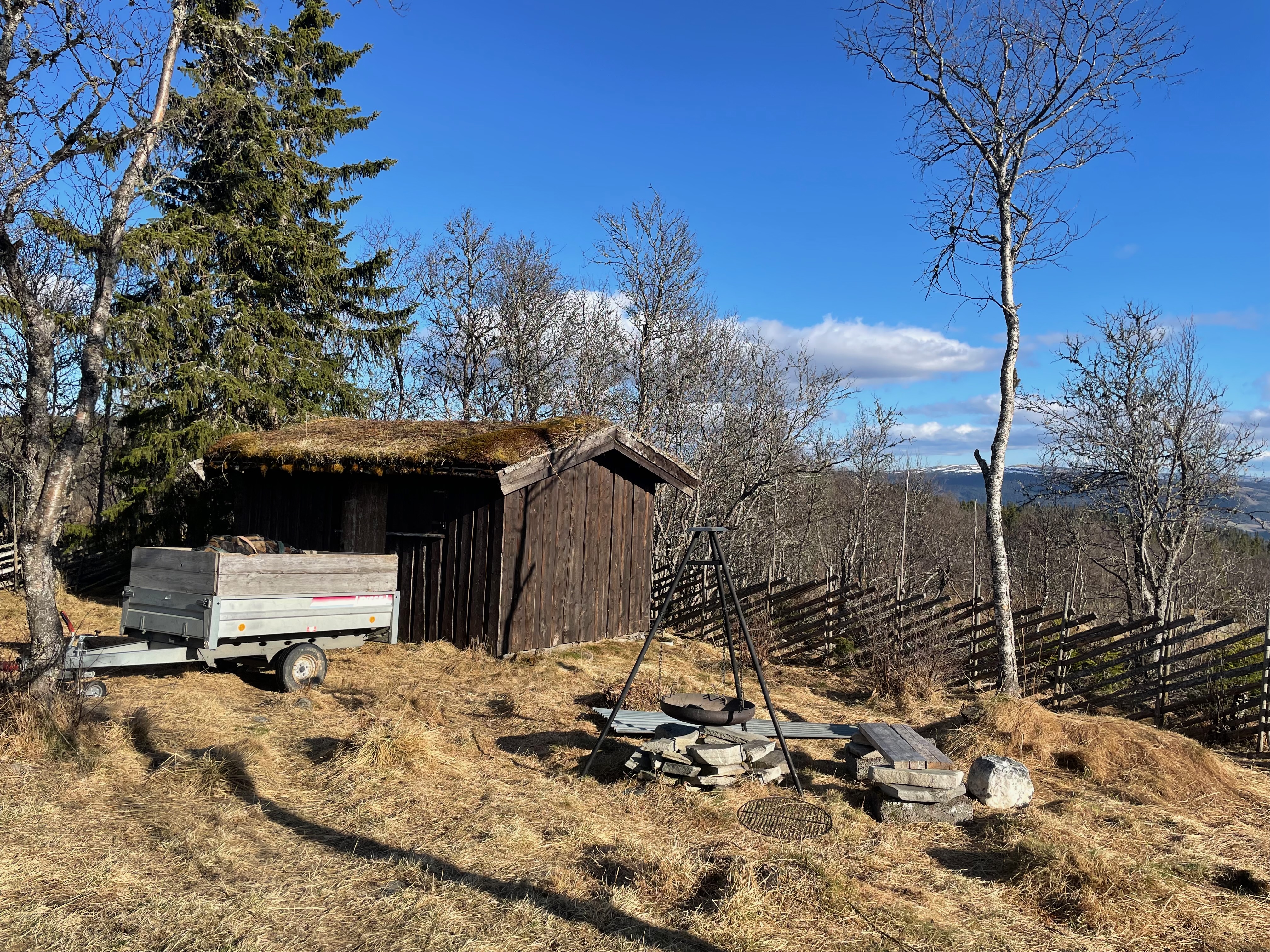 Per’s stuggu Sør-Fron outdoor fire pit with a black metal tripod grill, wooden turf roof shed, firewood trailer, dry mountain grass, bare trees and wide Norwegian valley views.