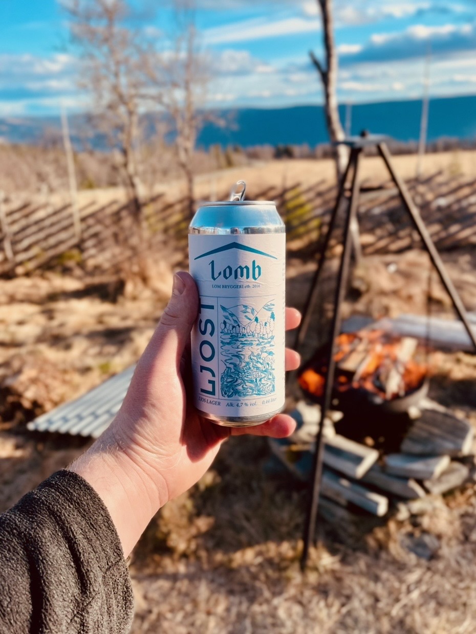 Per’s stuggu Sør-Fron outdoor fire pit with a hand holding Lom Bryggeri Ljos lager, black tripod grill, open flames, wooden fence and Norwegian mountain views.