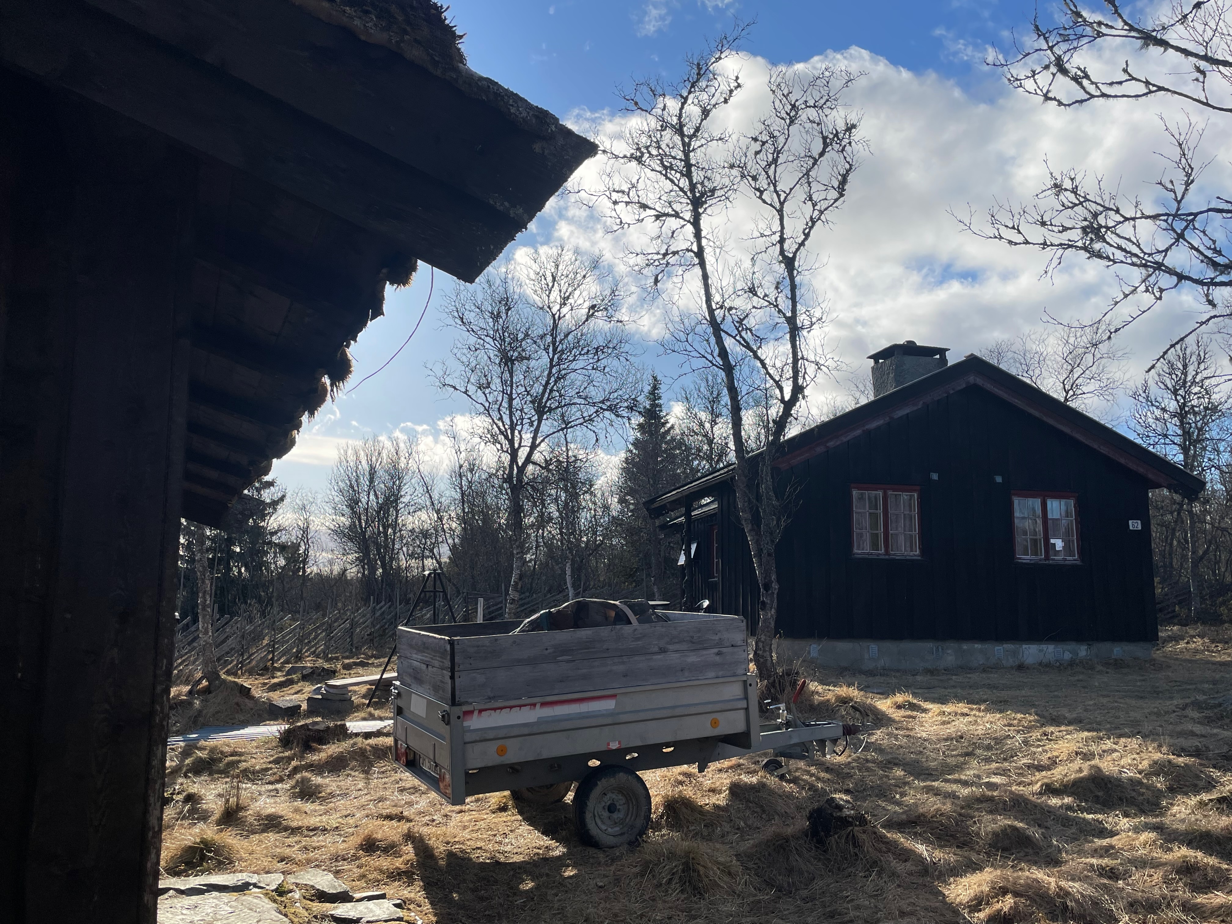 Per’s stuggu Sør-Fron rustic black wooden cabin with red window frames, firewood trailer, turf roof shed, bare trees and a dry mountain landscape in Norway.