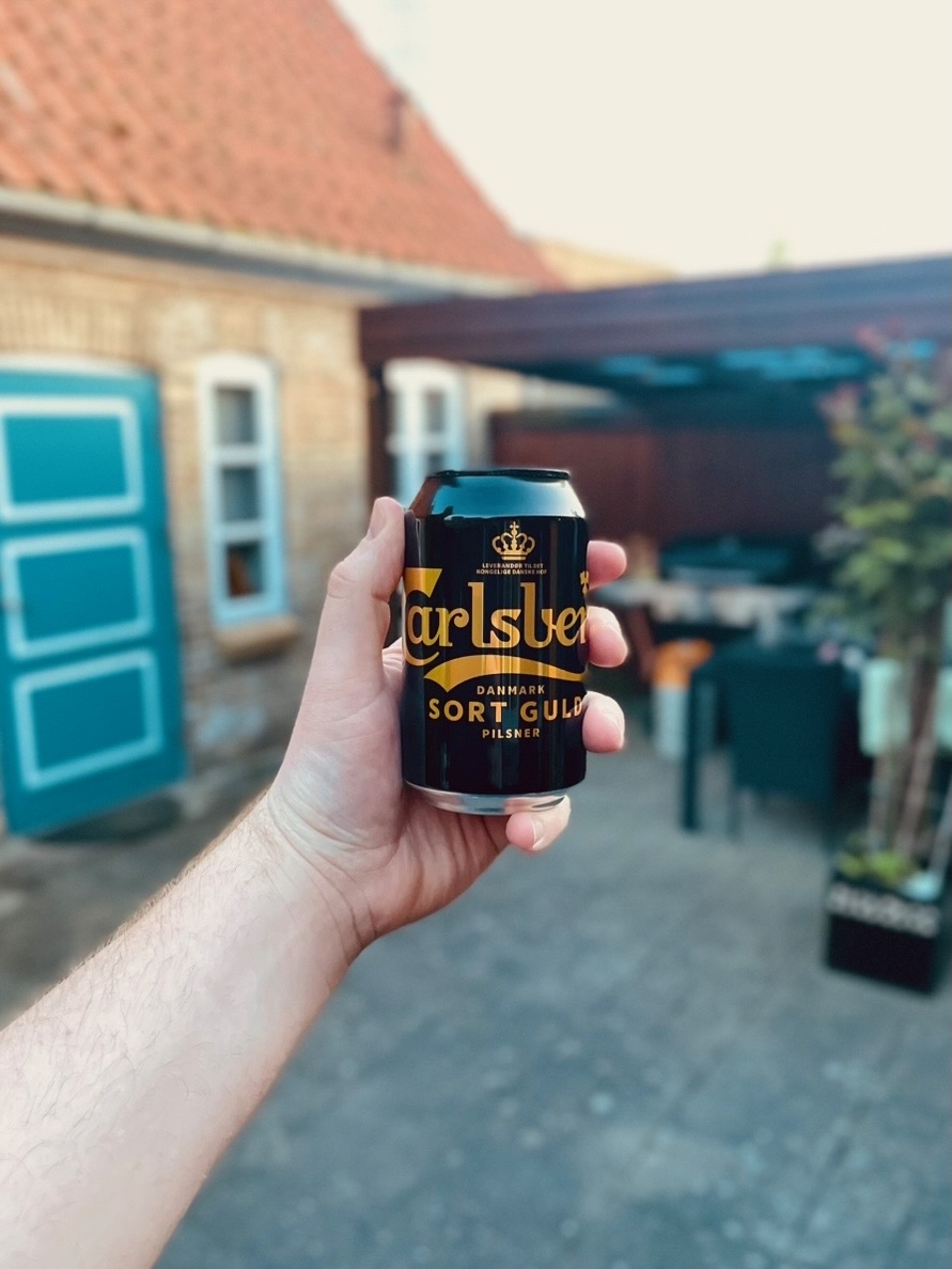 Private Annex Haderslev Review showing a hand holding a Carlsberg Sort Guld pilsner can in the cozy Airbnb courtyard in Haderslev, with the blue door and outdoor seating in the background.