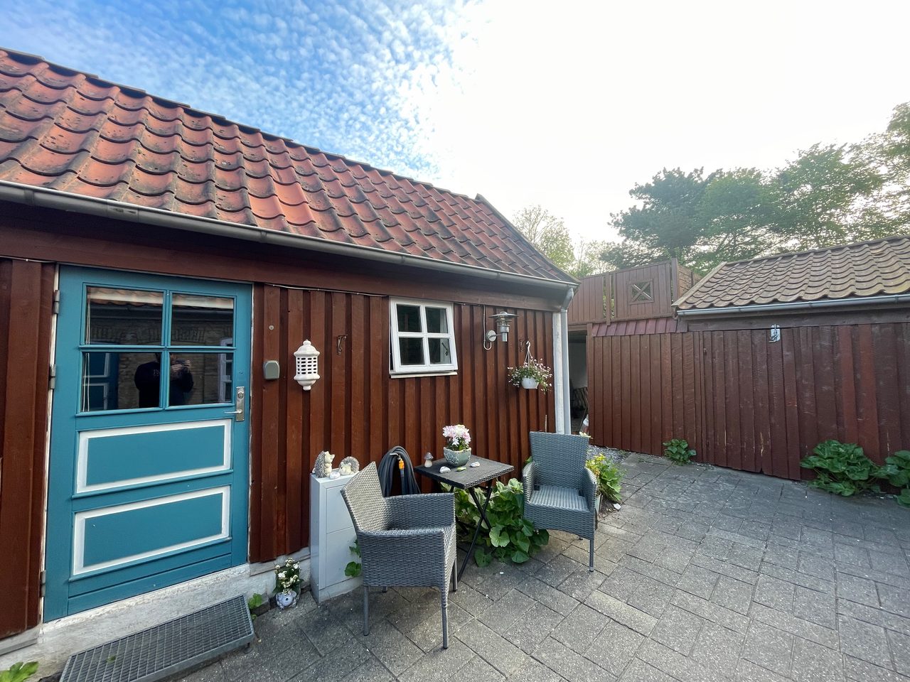 Private Annex Haderslev Review showing the cozy Airbnb courtyard in Haderslev with a blue door, wooden annex, outdoor chairs, small table, plants and red tiled roof.