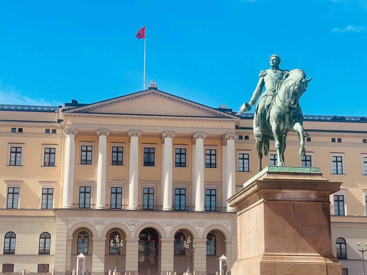 Royal Palace Oslo with the front facade of Det kongelige slott, white columns, the Norwegian flag, and the equestrian statue of King Karl Johan under a clear blue sky.
