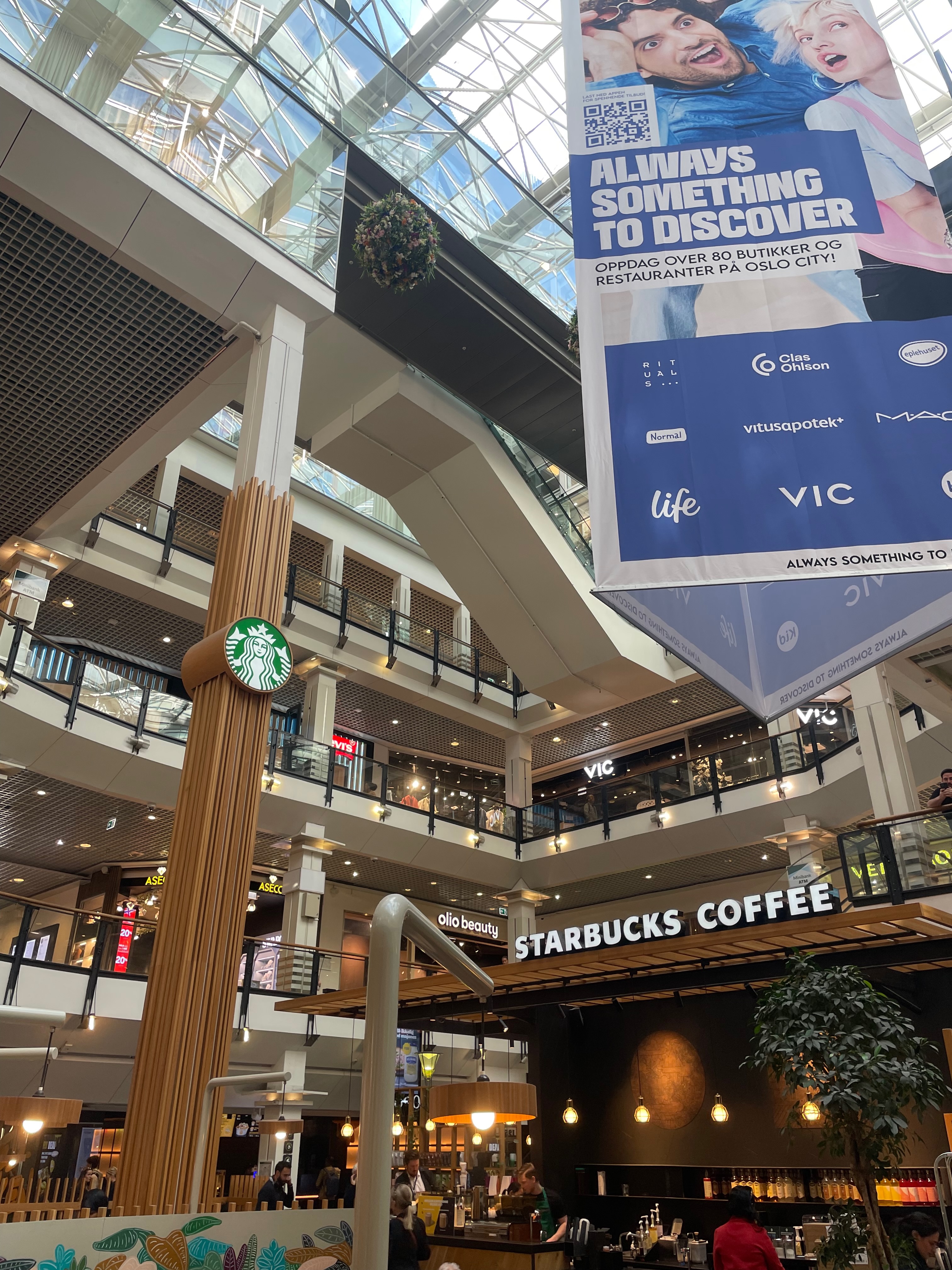 Starbucks Oslo City Review, Starbucks Coffee sign inside the Oslo City mall atrium with glass roof, hanging advertising banner, VIC store, wooden Starbucks pillar logo and café counter below