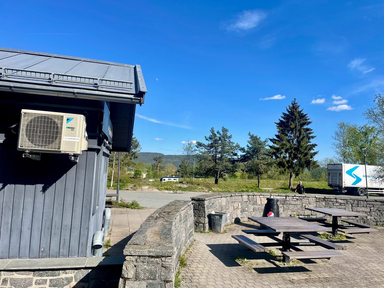Tollerud rasteplass syd outdoor seating area beside the E18 in Norway, with picnic tables, stone walls, parking space, trees, blue sky and the dark roadside restaurant building